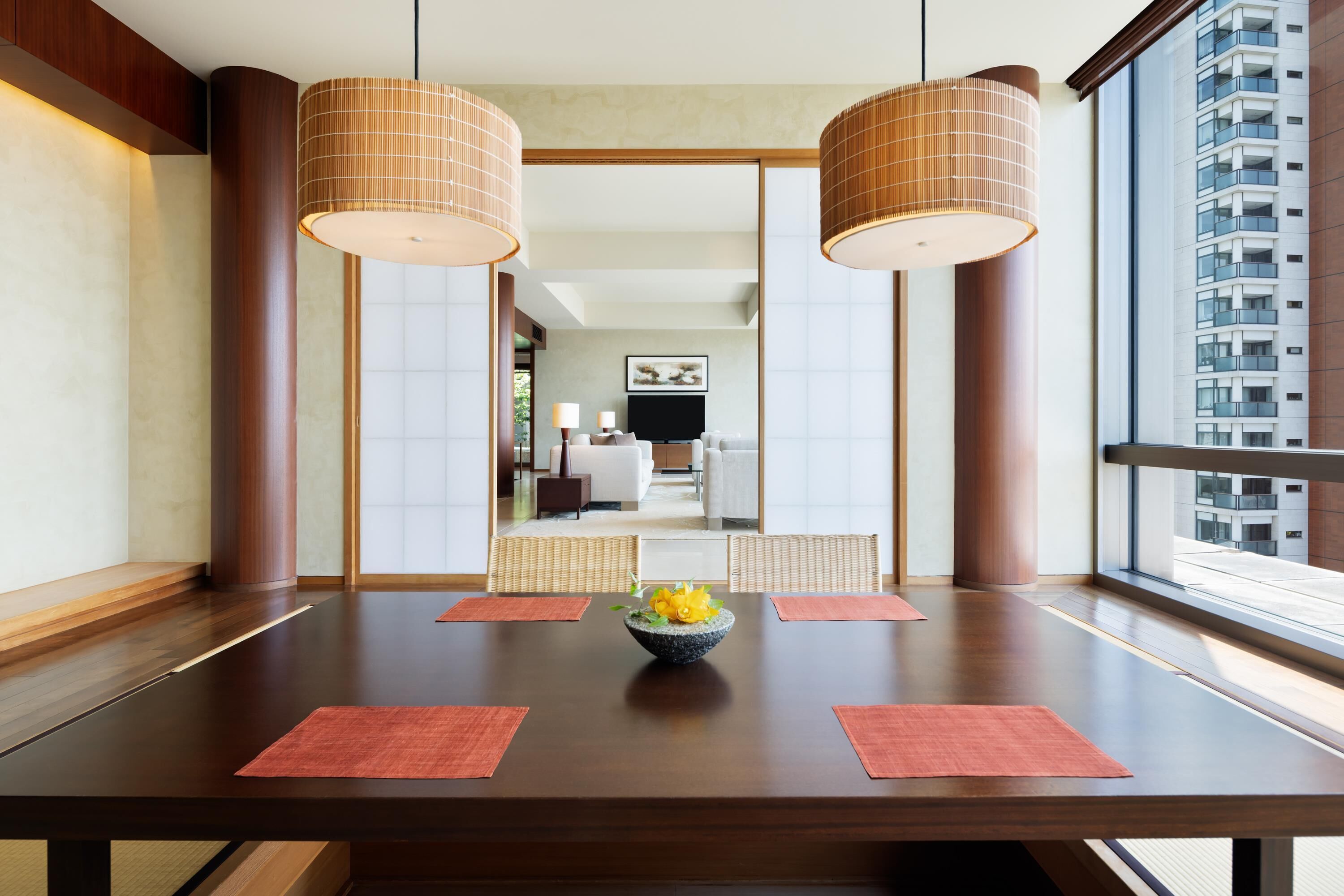 A modern hotel room with a wooden dining table, two large hanging lamps, and a view of the city through large windows.