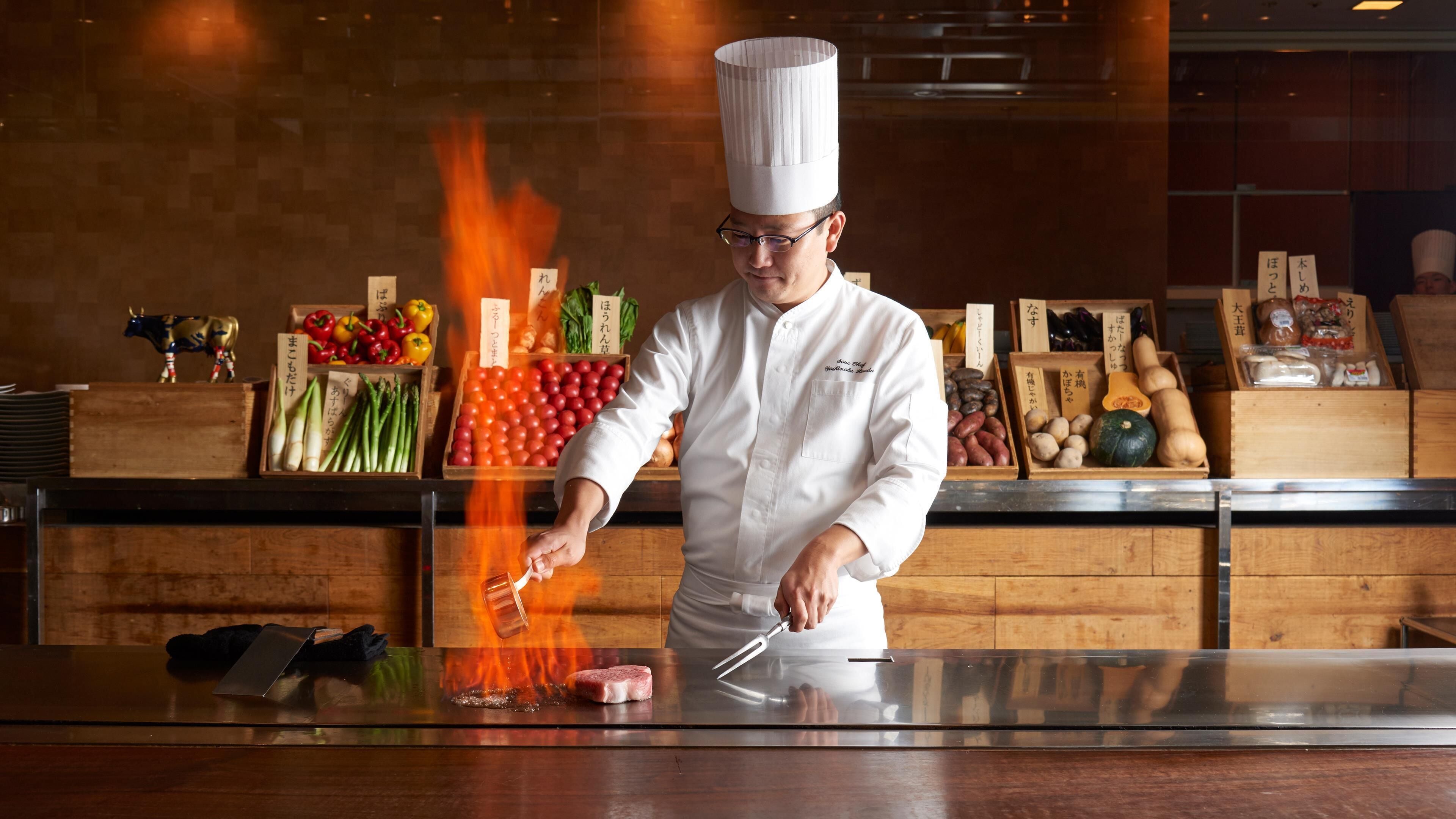 A chef performing a cooking technique with flames, surrounded by wooden crates of fresh produce and seafood.