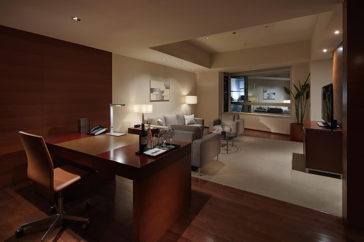A modern hotel room with a wooden desk, a sofa, a dining table, and a large window.