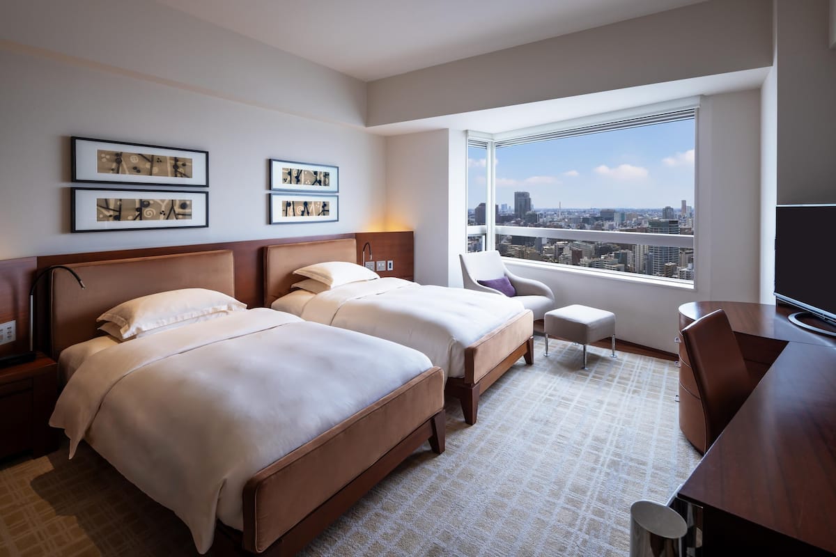 A hotel room with two beds, a desk, a chair, and a large window with a city view.