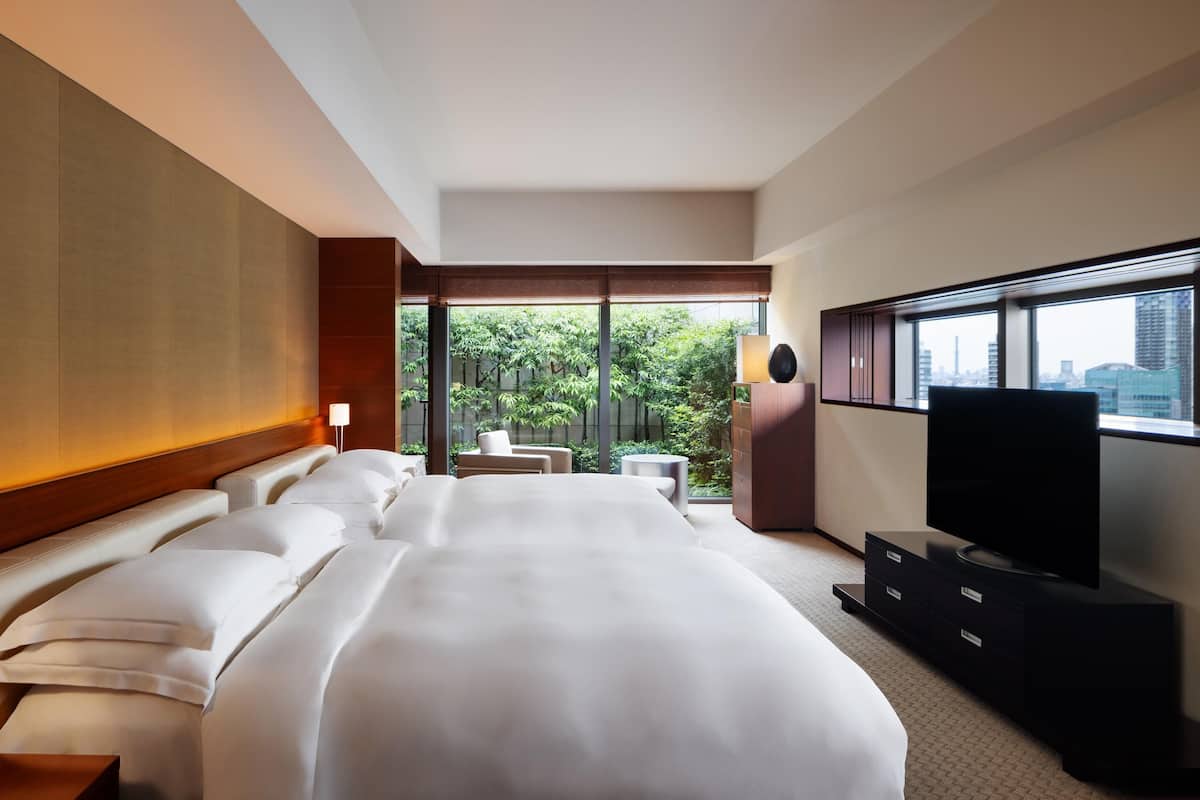 A hotel room with a large bed, a TV, a desk, and a view of greenery outside.