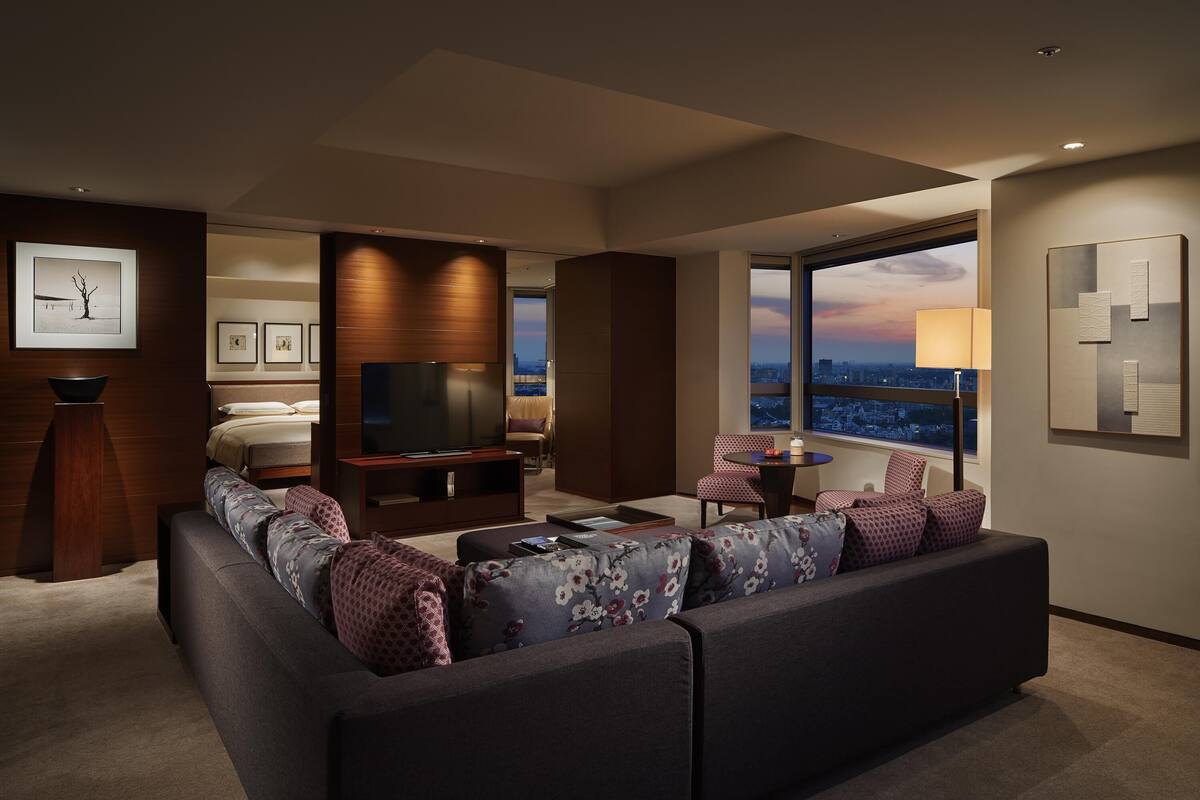 A modern hotel room with a large sofa, a TV, a desk, and a view of the cityscape.