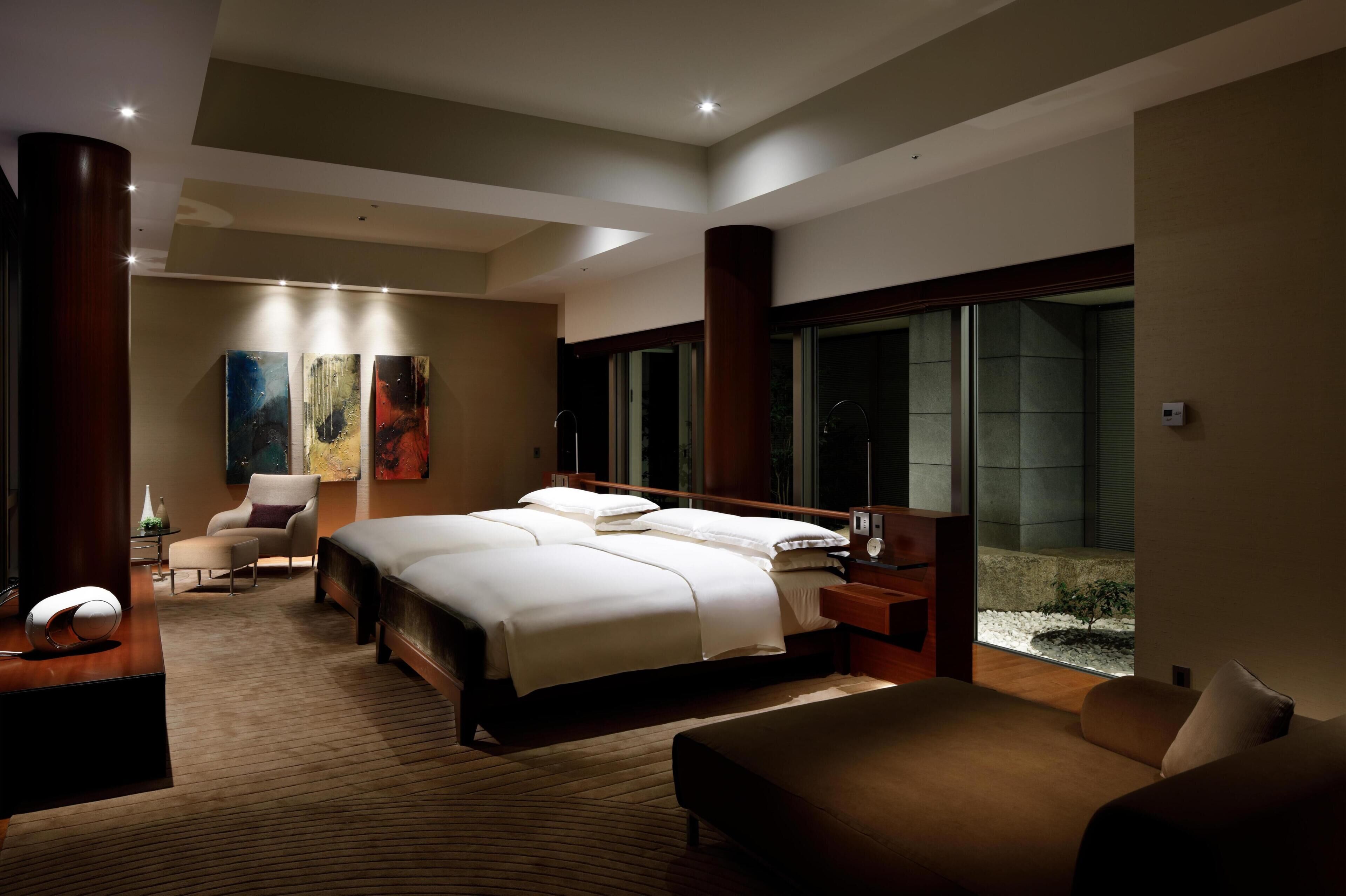 A modern hotel room with a large bed, a sitting area with a chair, a desk with a lamp, and a view of the outdoors through a large window.