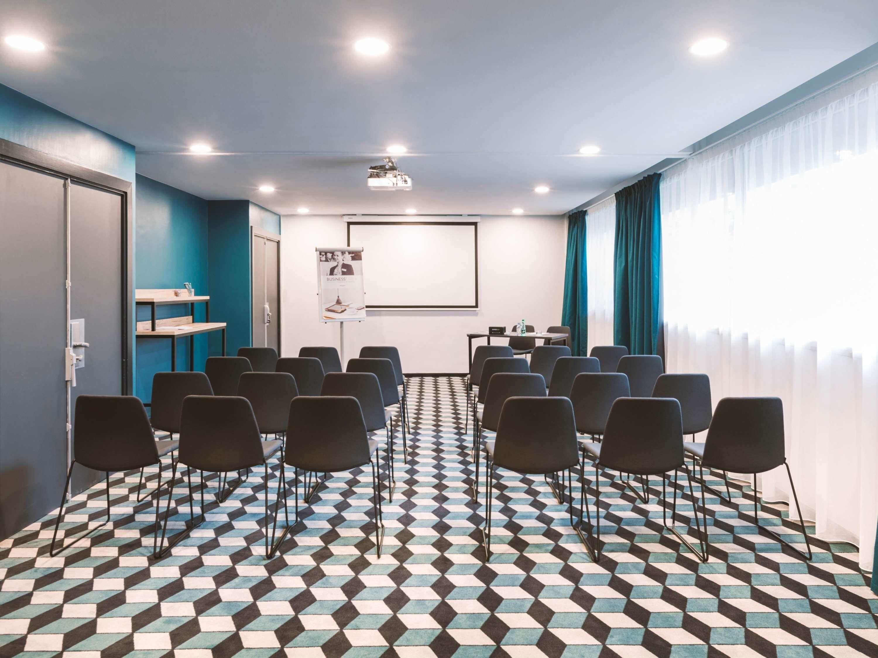 A conference room with a projector screen, black chairs, a patterned floor, and a window with curtains.