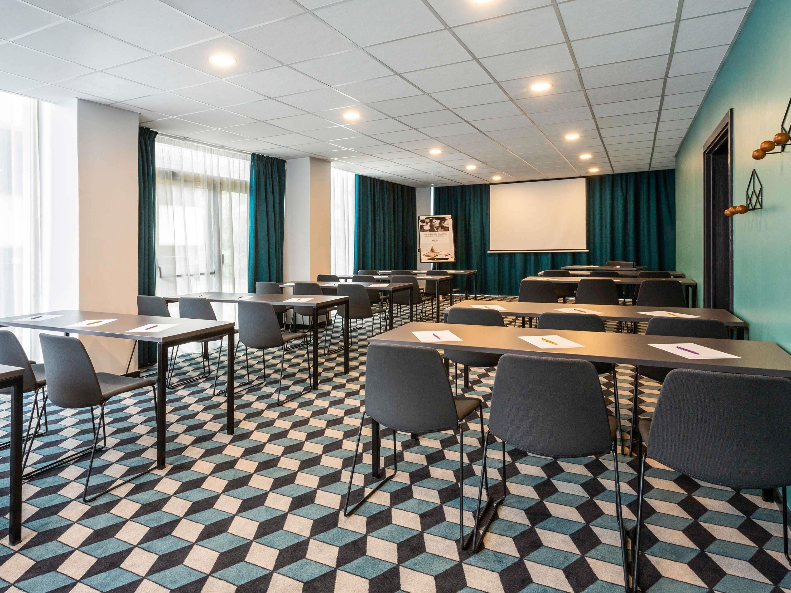 Une salle de conférence dotée d’un sol à carreaux, de tables avec des chaises, d’un écran de projection et d’une grande fenêtre avec des rideaux.