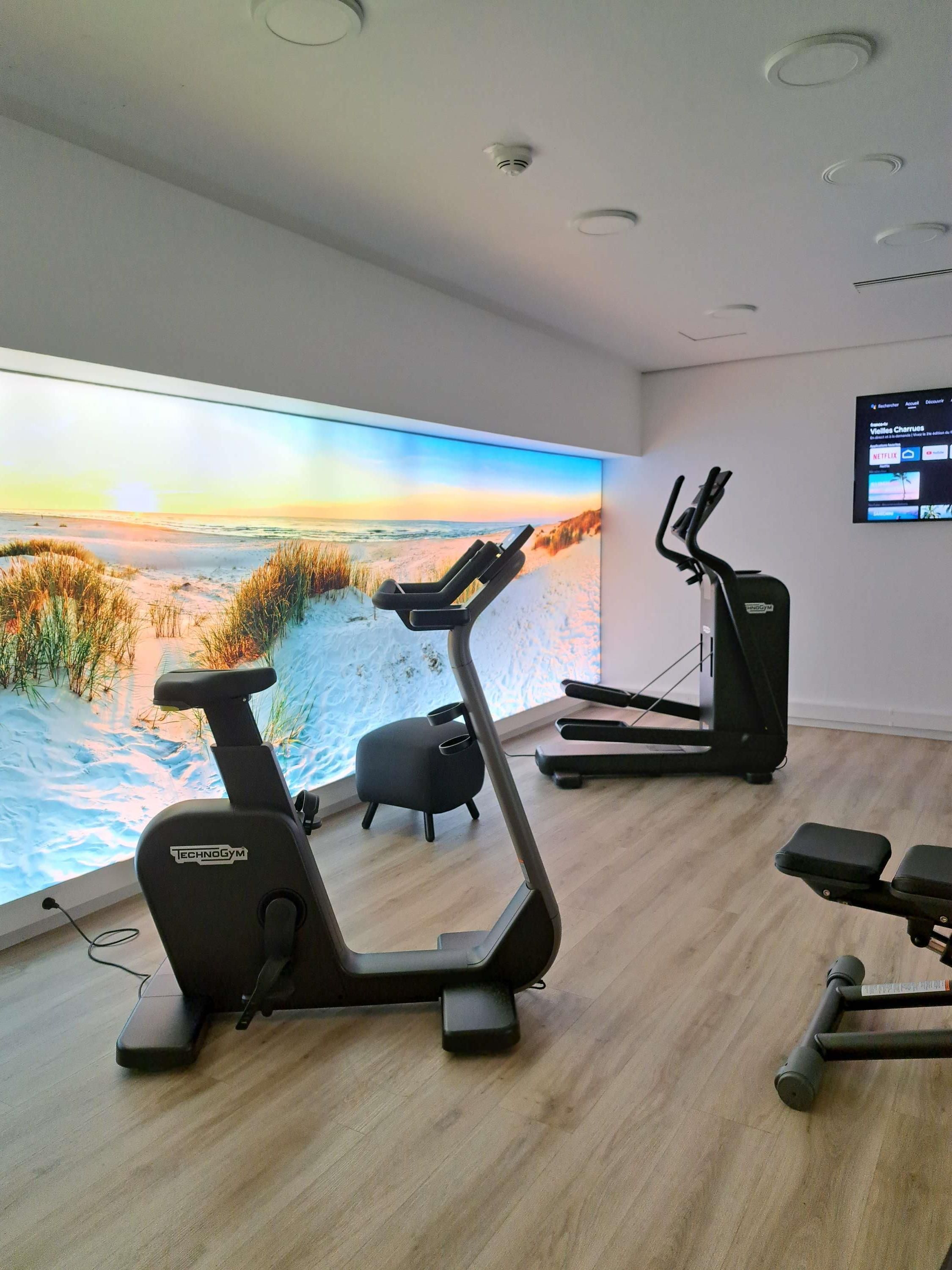 Une salle de sport équipée de matériel d’exercice, une grande fenêtre donnant sur la plage et un écran mural diffusant du contenu.