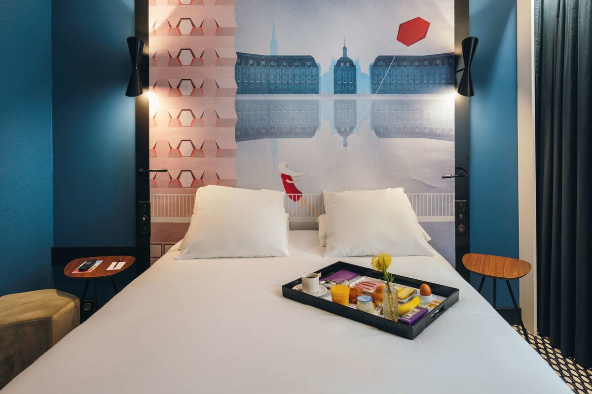 A hotel room with a large bed, two bedside tables, a tray with breakfast items, and a cityscape mural on the wall.
