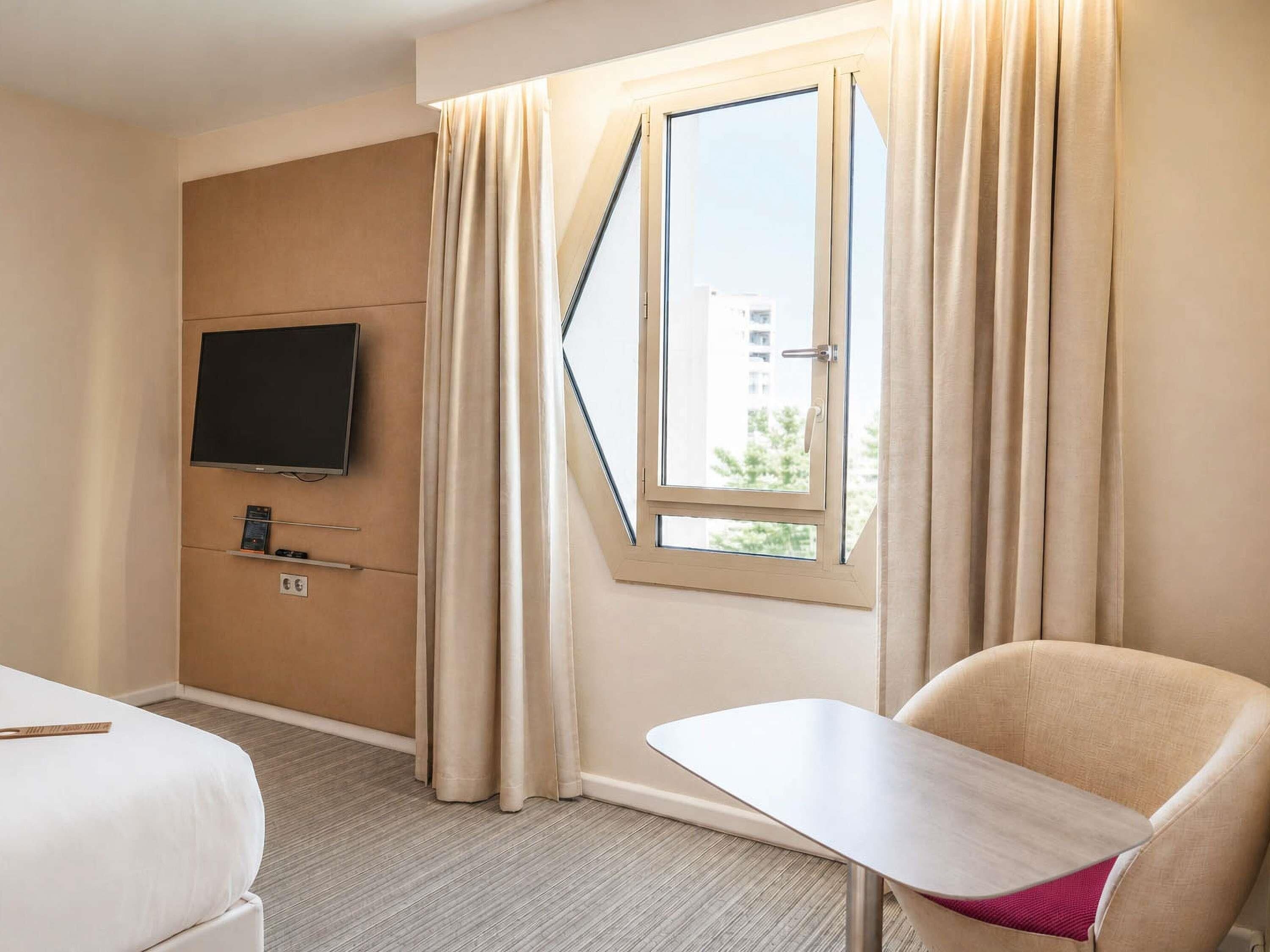 A hotel room with a bed, a TV mounted on the wall, a window with curtains, and a small table.