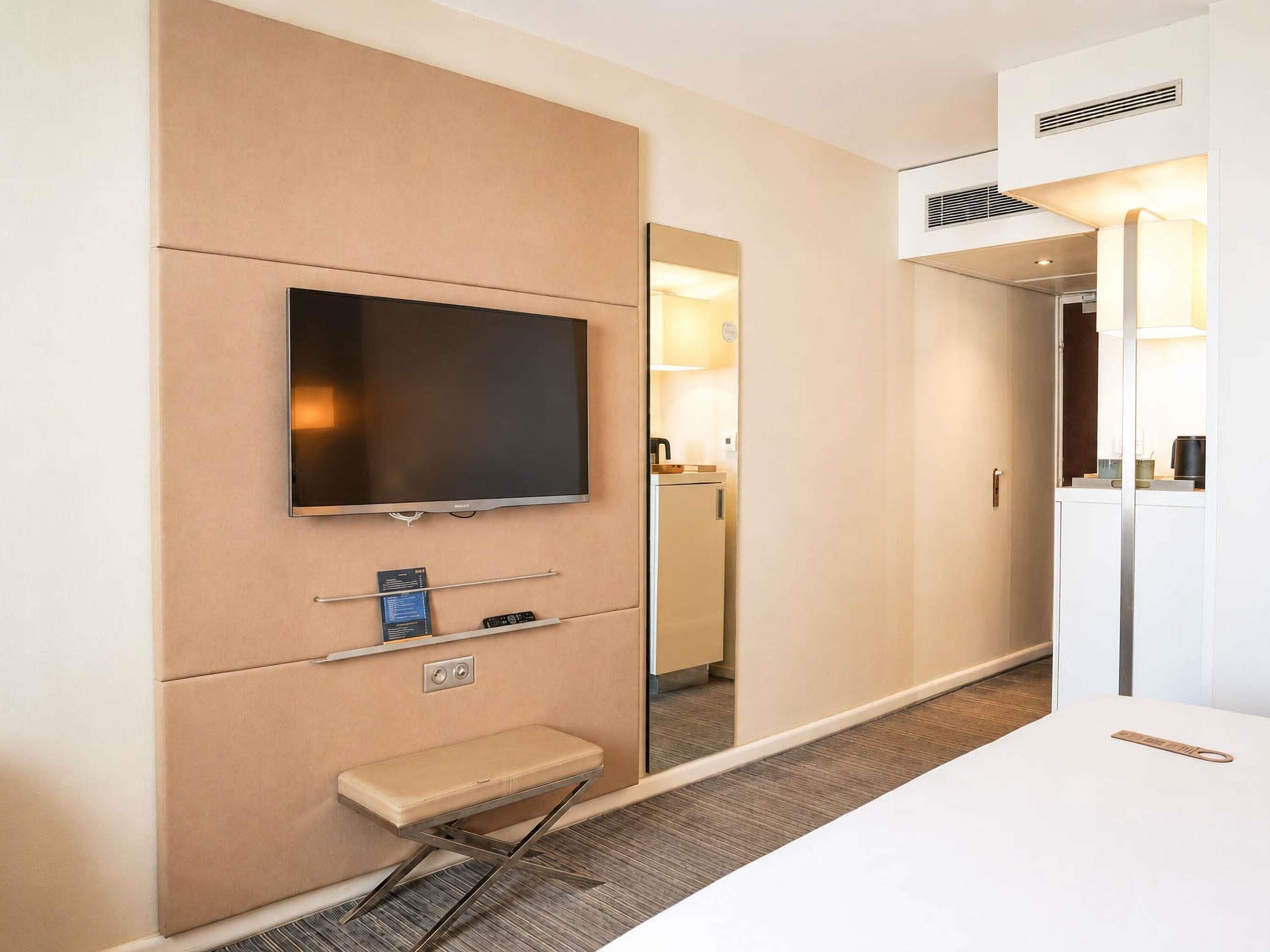 Ein Hotelzimmer mit Flachbildfernseher an der Wand, einem kleinen Nachttisch, einer Bank und einem Spiegel.