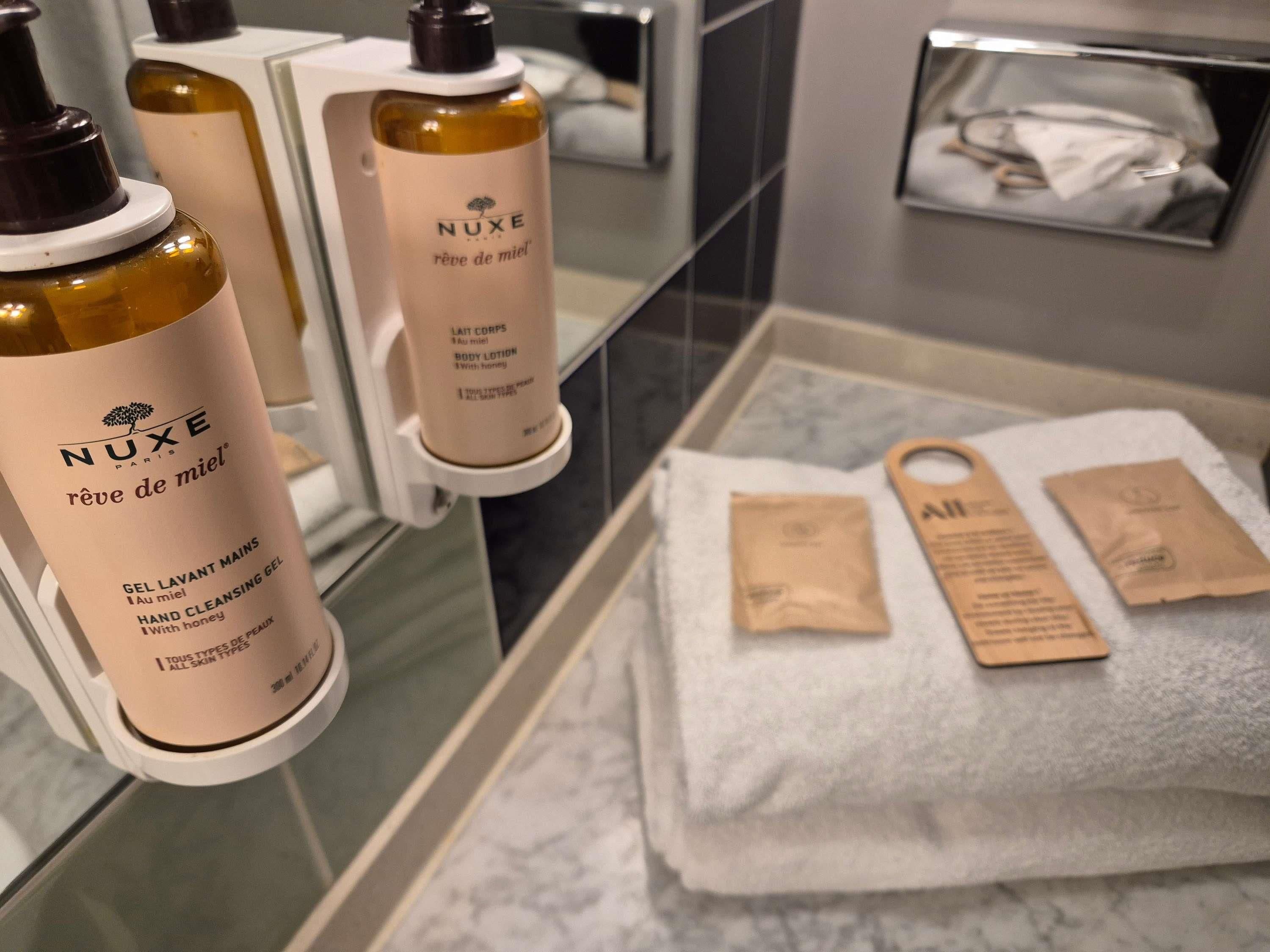 Un baño con loción corporal Nuxe y gel limpiador de manos, una toalla y una pequeña bolsa de madera.