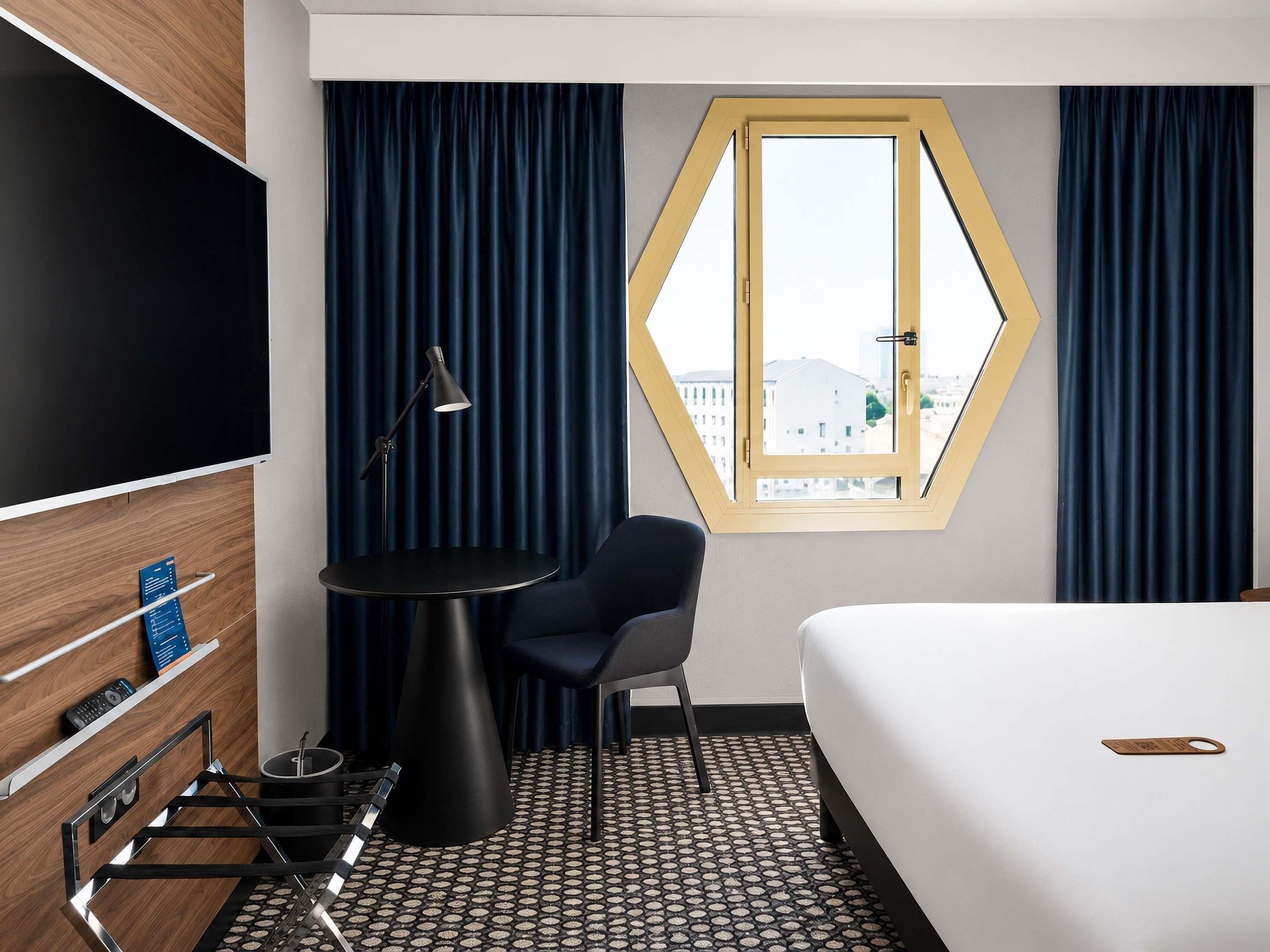 Habitación de hotel con cama, escritorio con silla, televisor, ventana con cortinas y vista a edificios.