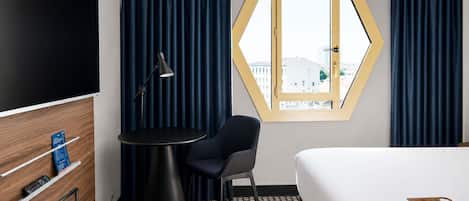 Ett hotellrum med en säng, ett skrivbord med en stol, en tv, ett fönster med gardiner och utsikt över byggnader.