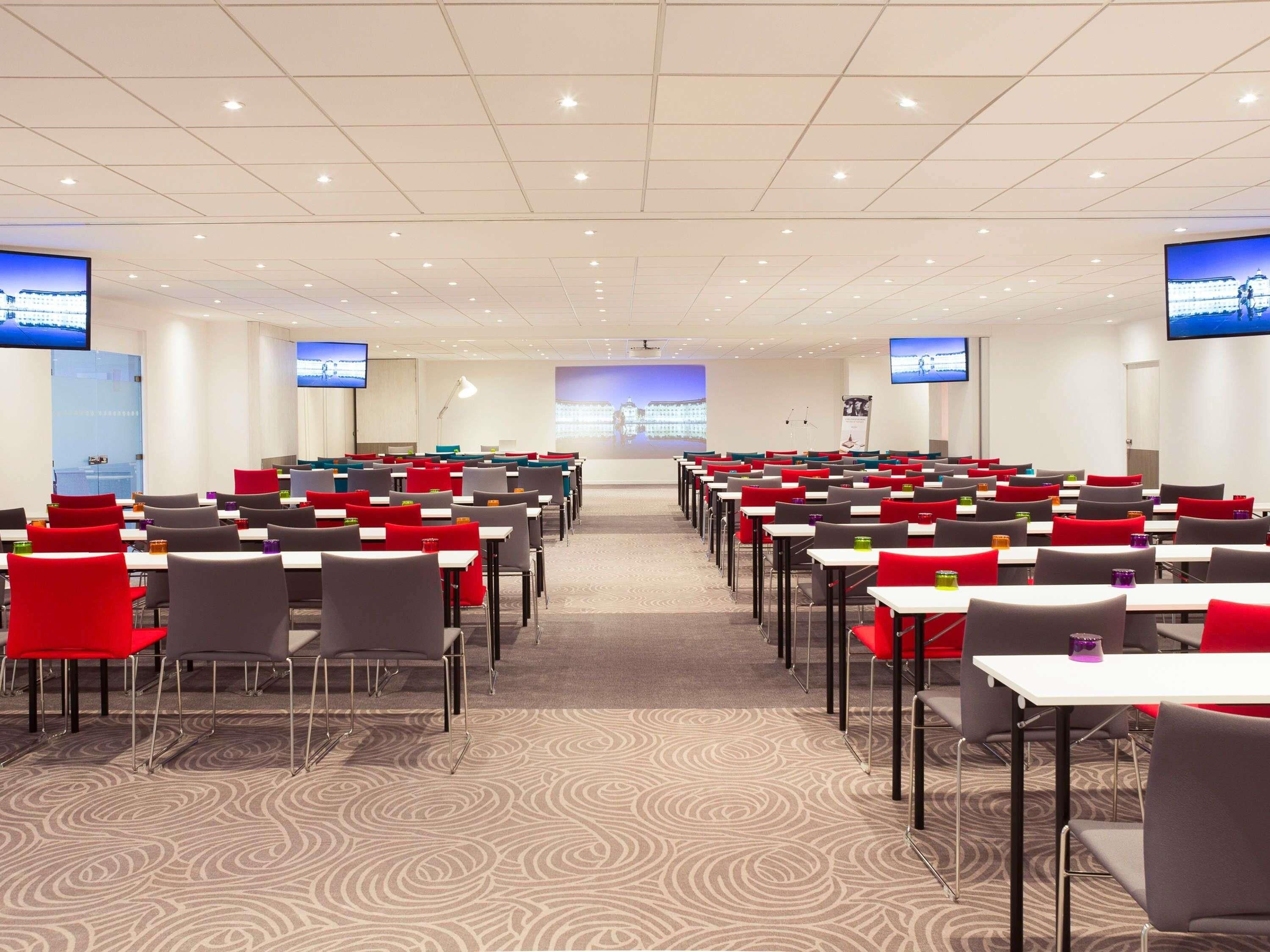Une salle de conférence avec des rangées de tables et de chaises, équipée de téléviseurs à écran plat et d’un grand écran situé au fond de la salle.