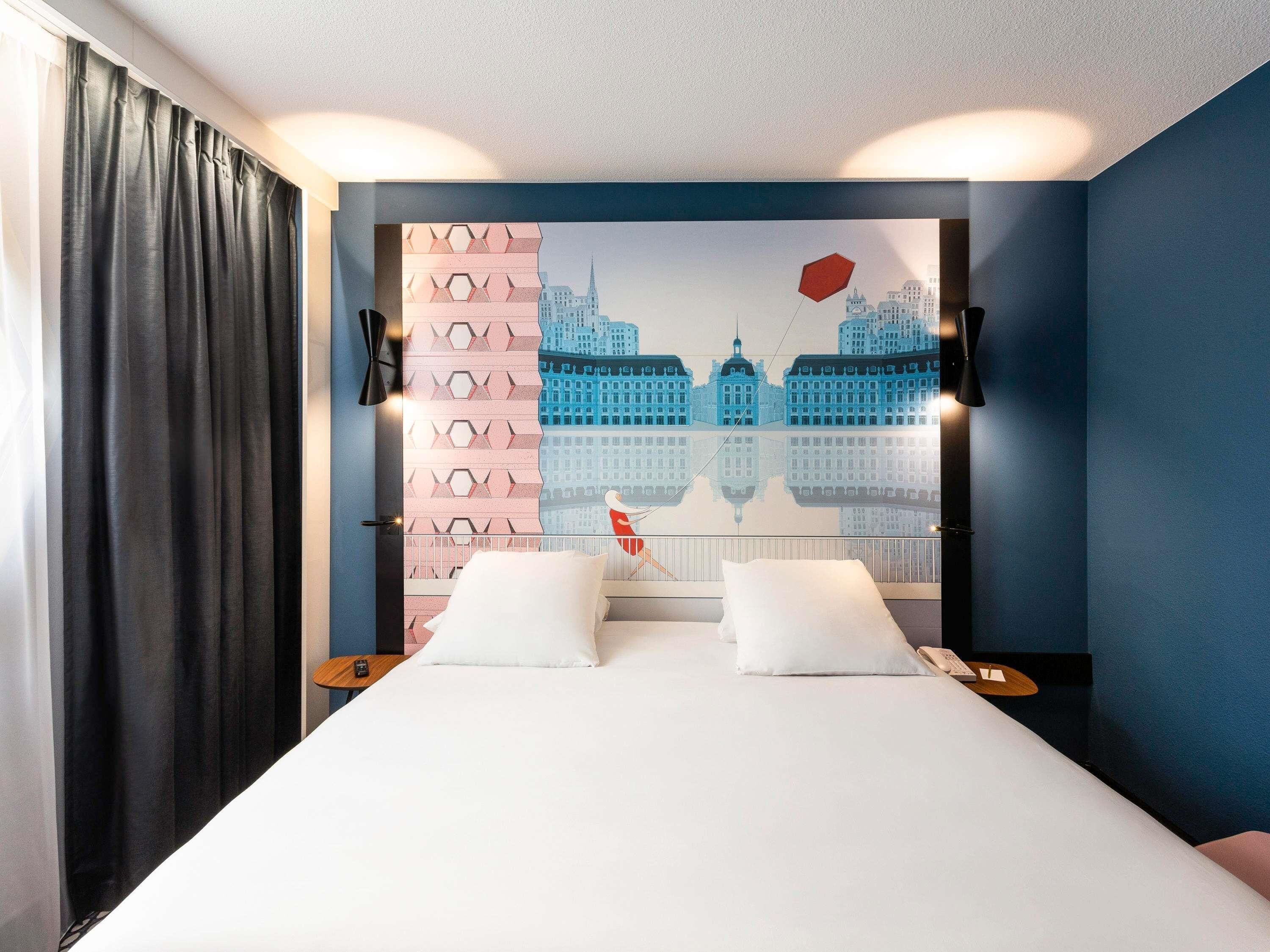 A hotel room with a bed, two bedside tables, a nightstand, a wall-mounted lamp, and a cityscape mural.