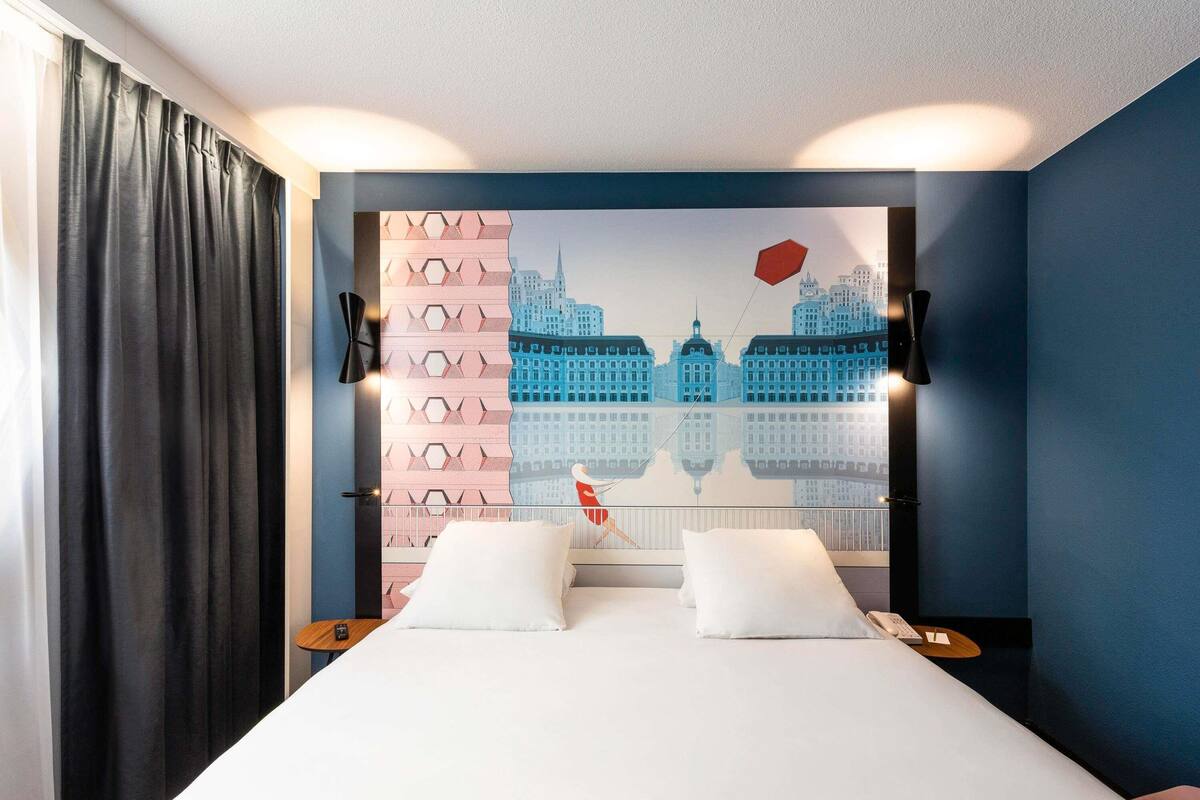 A hotel room with a bed, two bedside tables, a nightstand, a wall-mounted lamp, and a cityscape mural.