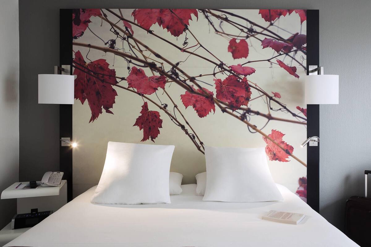 A hotel room with a bed, two bedside lamps, a nightstand with a phone, and a wall mural of red leaves.
