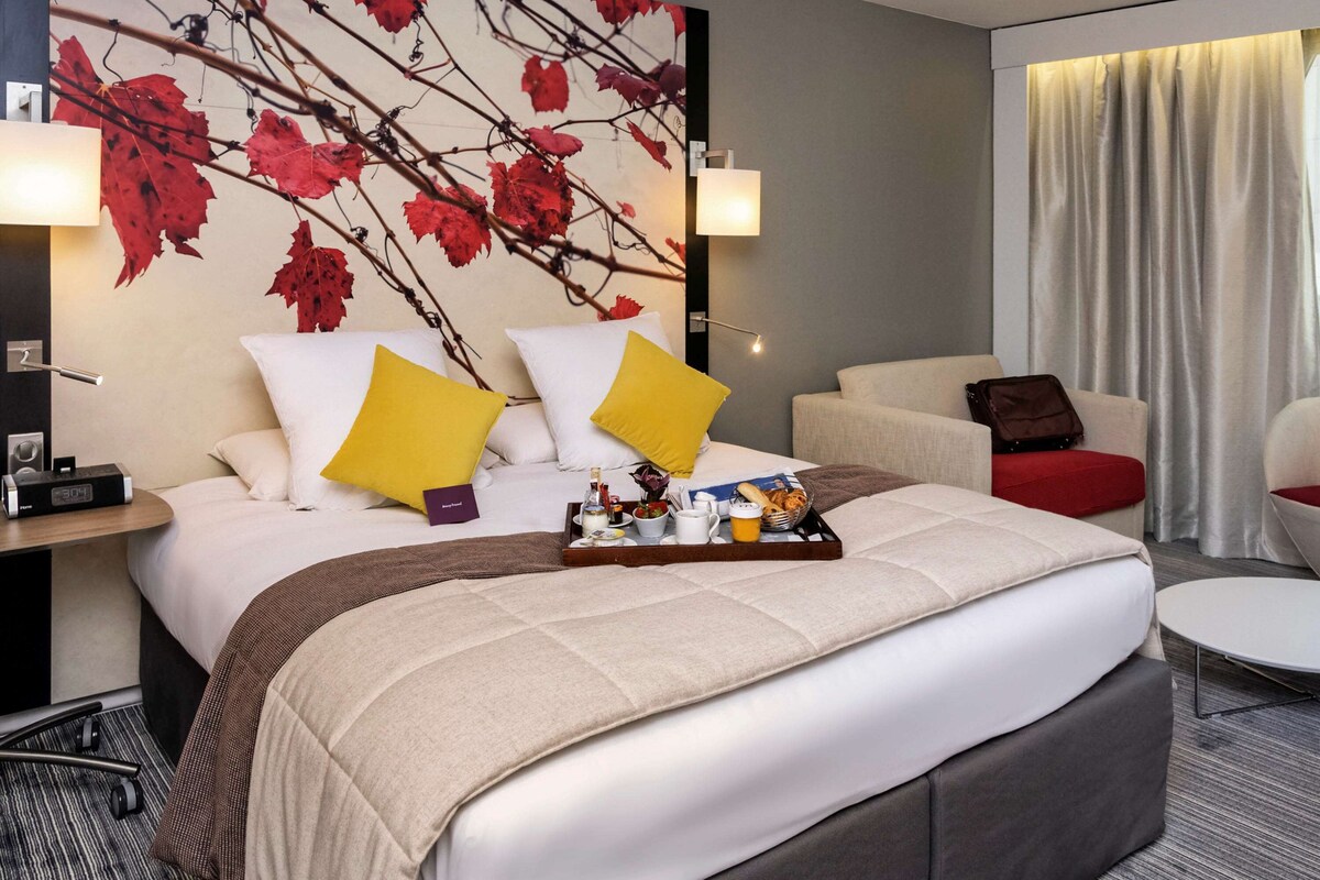 A hotel room with a large bed, two bedside tables, a desk, a chair, a small table with breakfast items, and a wall with a leaf pattern.