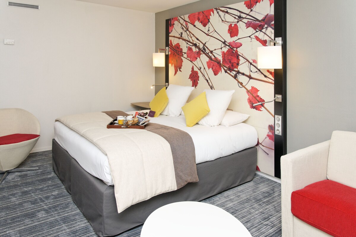 A hotel room with a bed, a chair, a small table, and a wall with a leaf pattern.