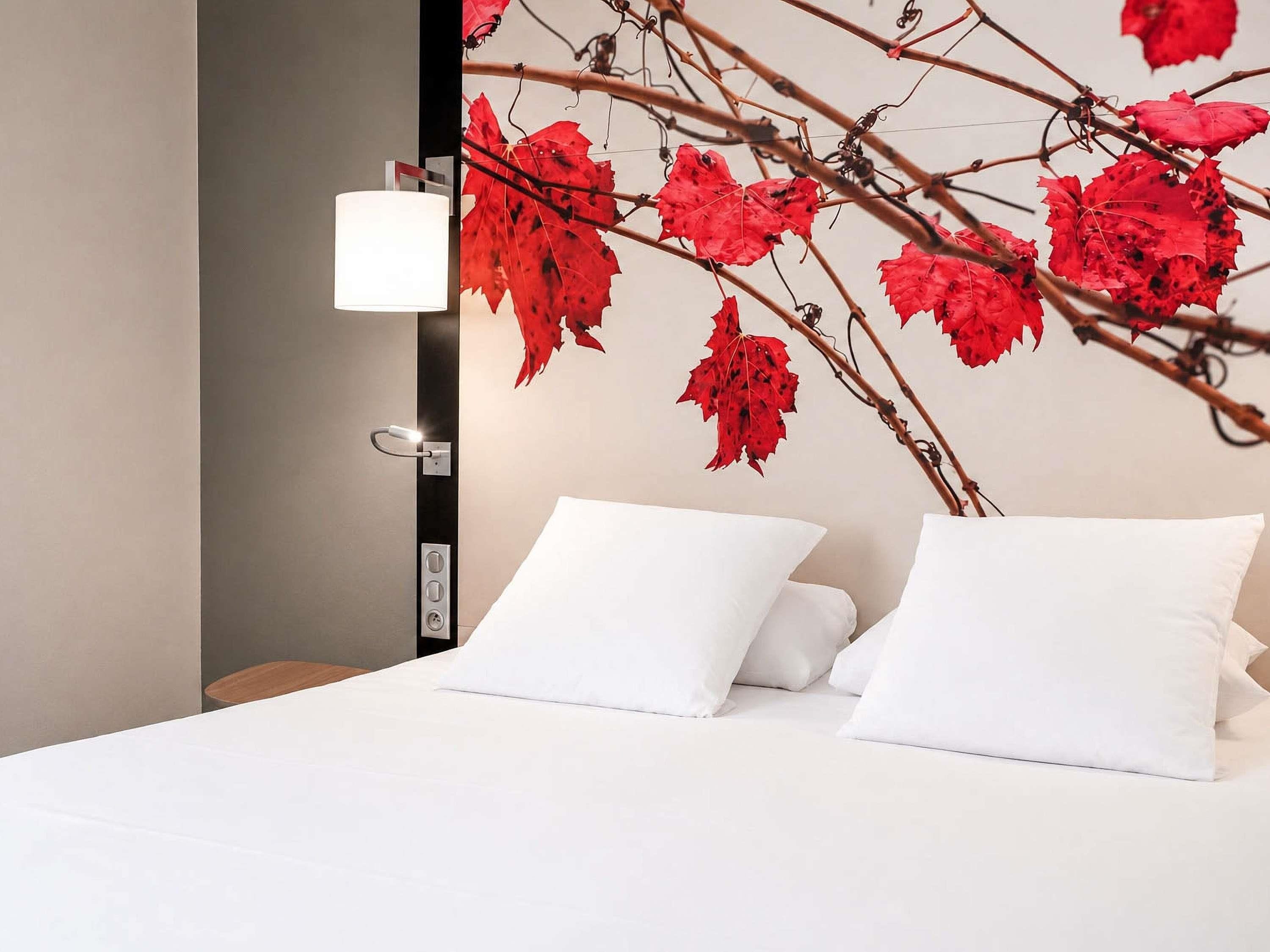 A hotel room with a bed, white pillows, a wall-mounted lamp, and a branch with red leaves.