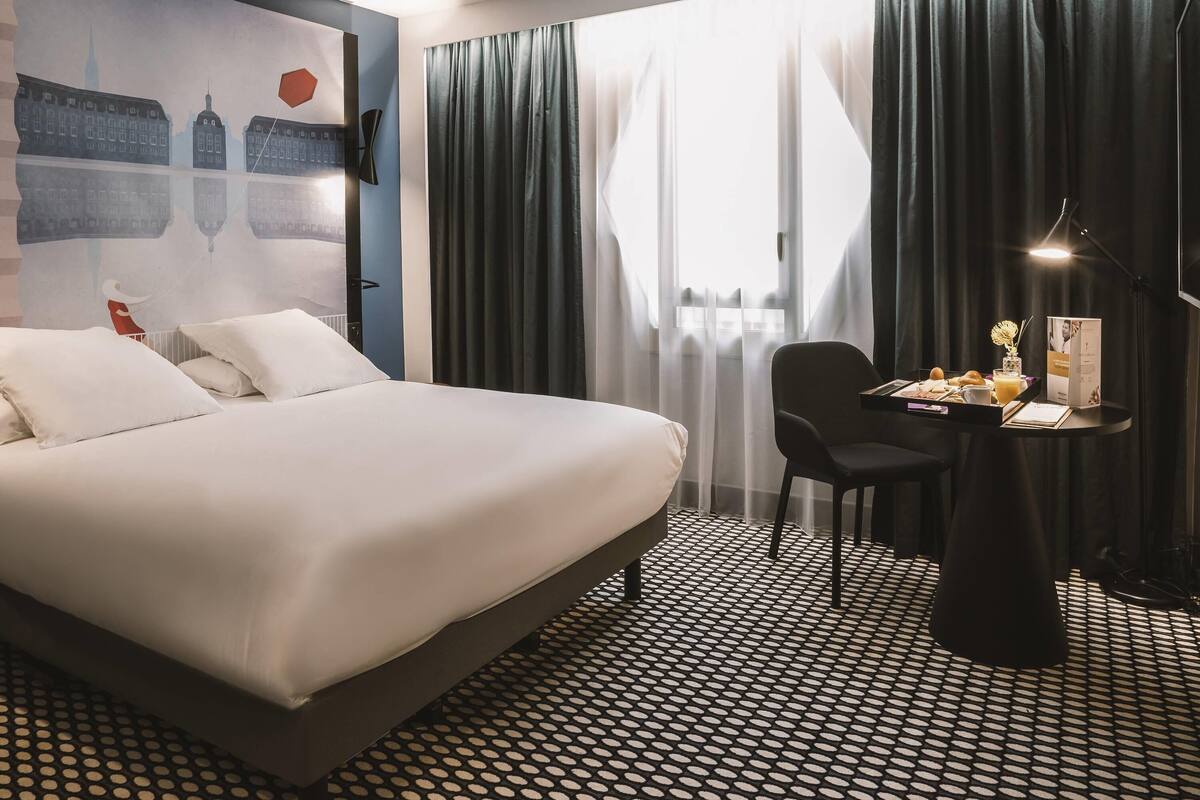 A hotel room with a large bed, a desk with breakfast, and a view of the city through sheer curtains.