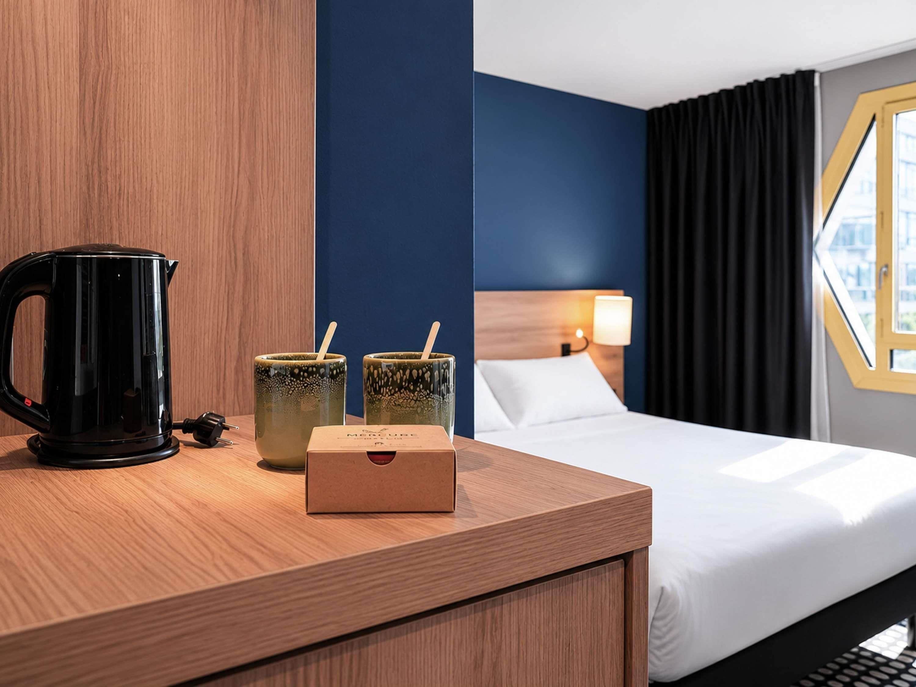 Una habitación de hotel con una mesita de noche de madera, un hervidor eléctrico negro, dos tazas de cerámica y una caja con el rótulo 'MERCURE'.