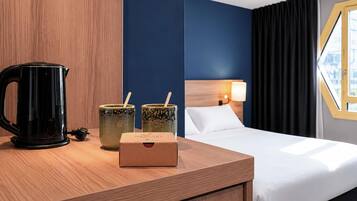 Una habitación de hotel con una mesita de noche de madera, un hervidor eléctrico negro, dos tazas de cerámica y una caja con el rótulo 'MERCURE'.