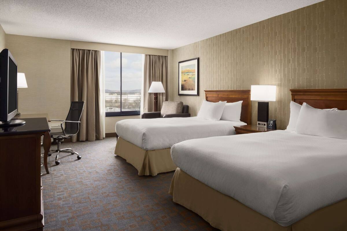 A hotel room with two beds, a desk with a computer, a chair, and a window with a view of the city.