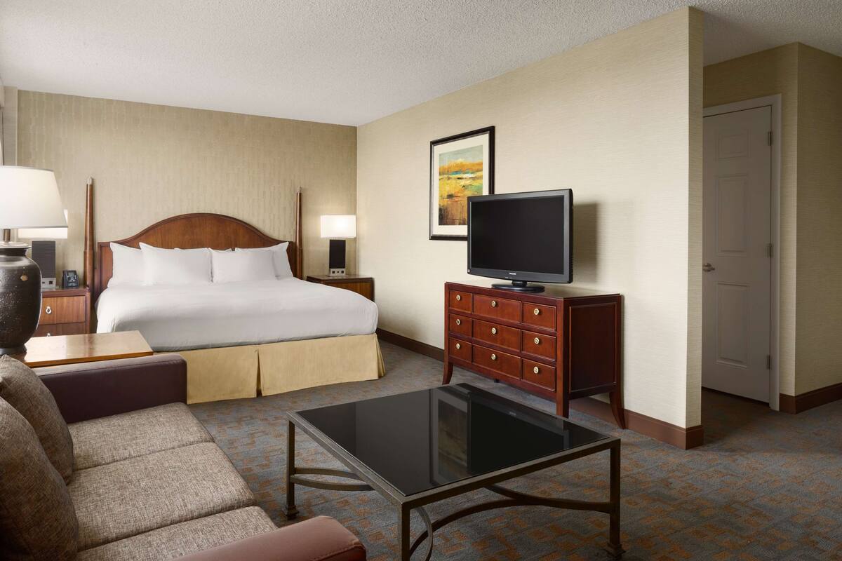 A hotel room with a bed, a television on a wooden dresser, a coffee table, a sofa, and a nightstand with lamps.