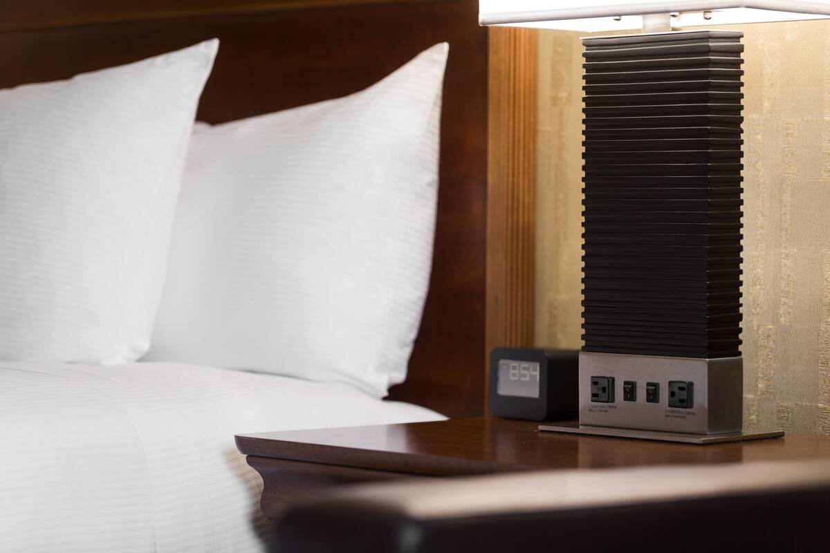 A neatly made bed with white linens, a bedside table with a black clock, and a wall-mounted air conditioning unit.