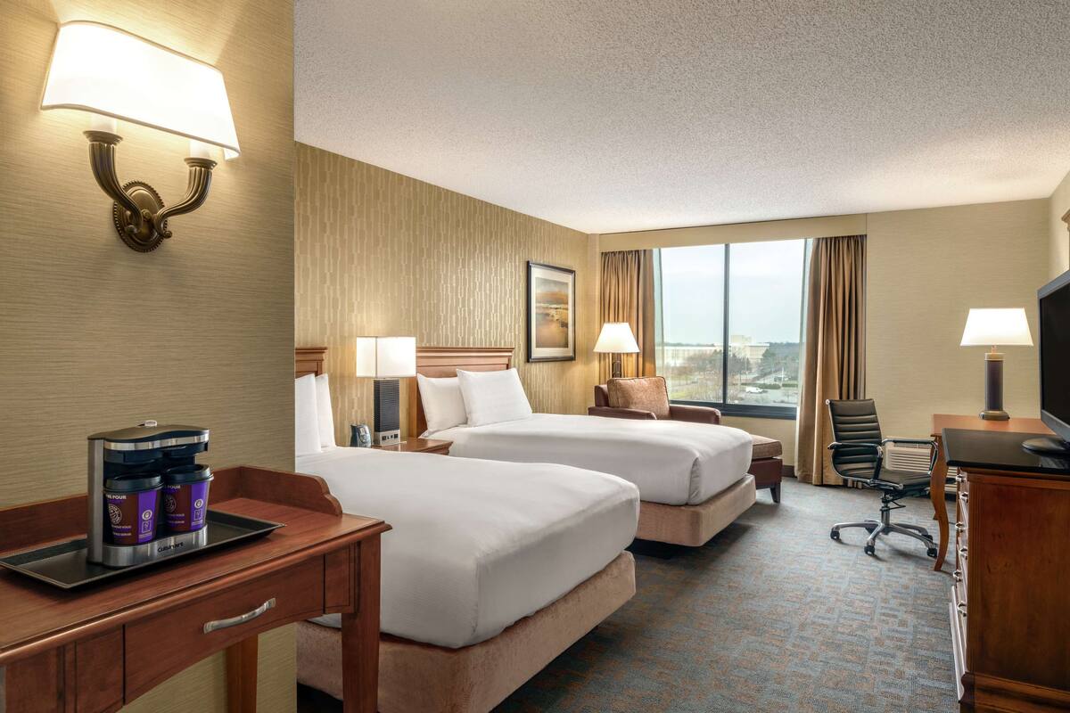 A hotel room with two beds, a desk with a coffee maker, a TV, and a large window with a city view.