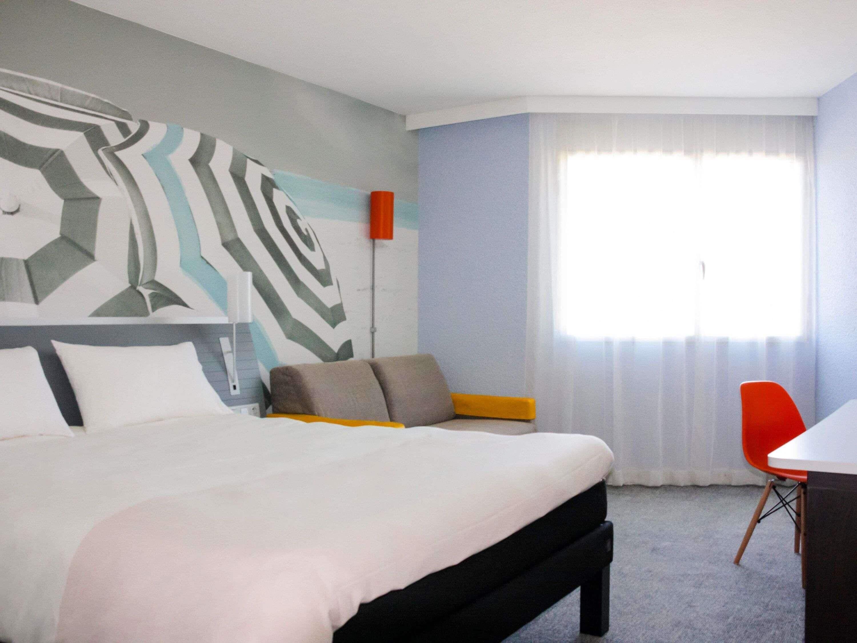 Una habitación de hotel moderna con una cama, un escritorio, una silla y un ventanal con cortinas.