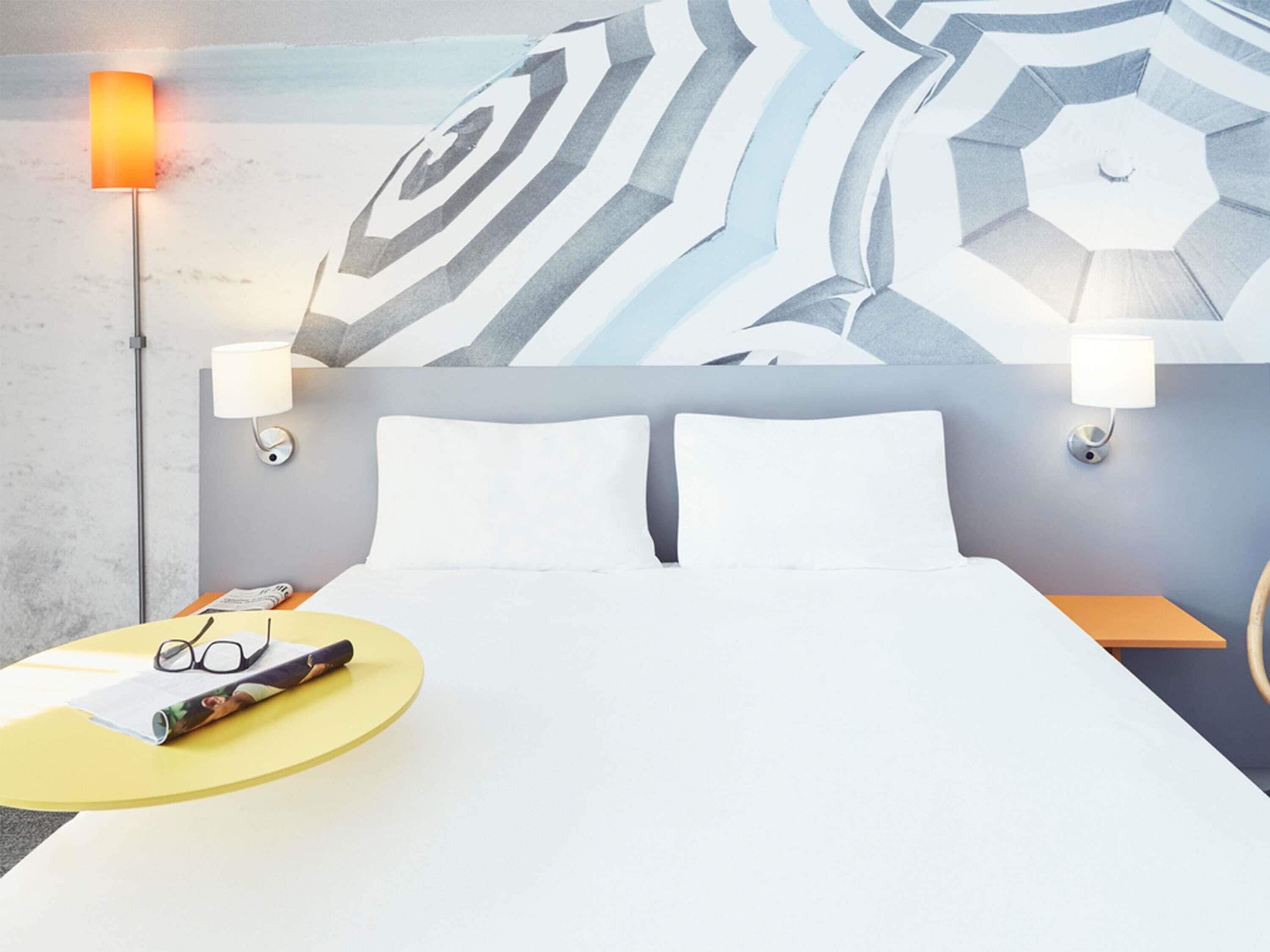 A hotel room with a bed, two bedside lamps, a wall with a geometric pattern, a round yellow table with glasses and a magazine, and a nightstand with a lamp.