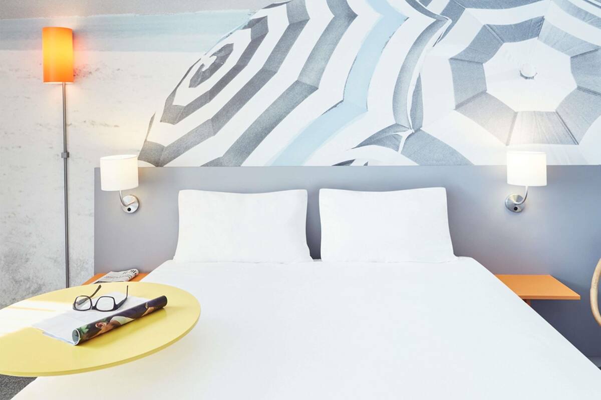 Une chambre d’hôtel avec un lit, deux lampes de chevet, un mur orné d’un motif géométrique, une table ronde jaune avec des verres et un magazine, et une table de chevet avec une lampe.