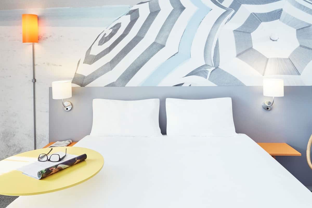 A hotel room with a bed, two bedside lamps, a wall with a geometric pattern, a round yellow table with glasses and a magazine, and a nightstand with a lamp.