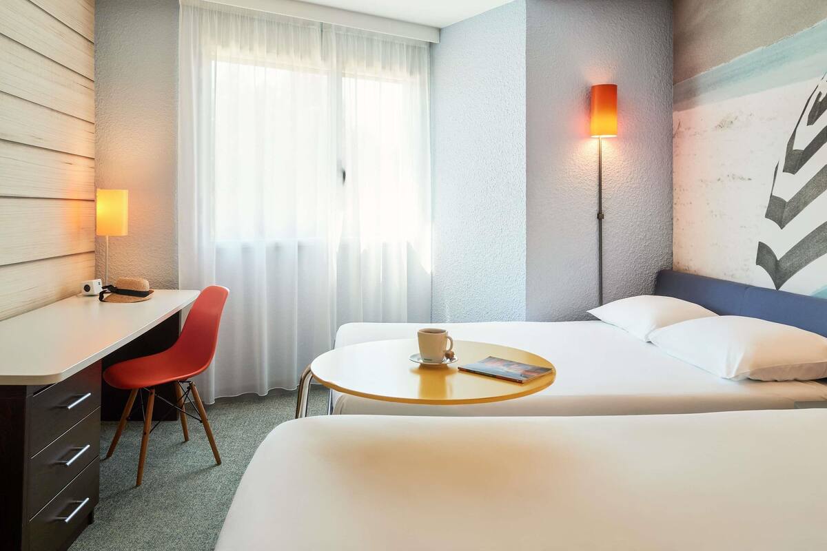 Une chambre d’hôtel équipée d’un bureau, d’une chaise, d’un lit, d’une table ronde avec une télécommande et d’une lampe fixée au mur.