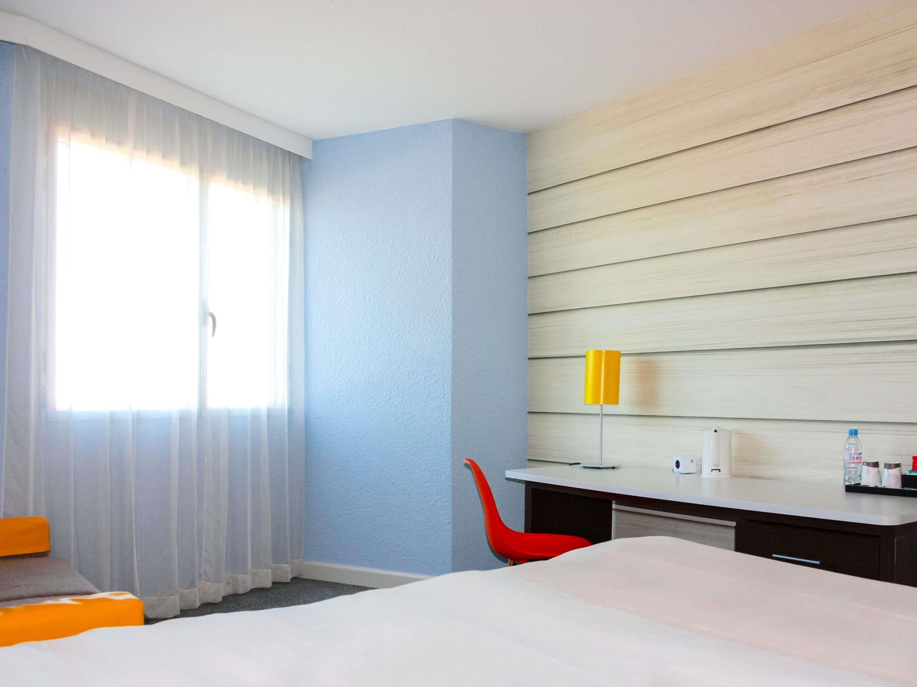 A hotel room with a bed, a desk, a chair, a window with curtains, and a wall with horizontal wooden panels.