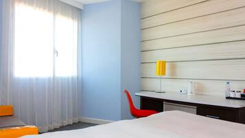 A hotel room with a bed, a desk, a chair, a window with curtains, and a wall with horizontal wooden panels.