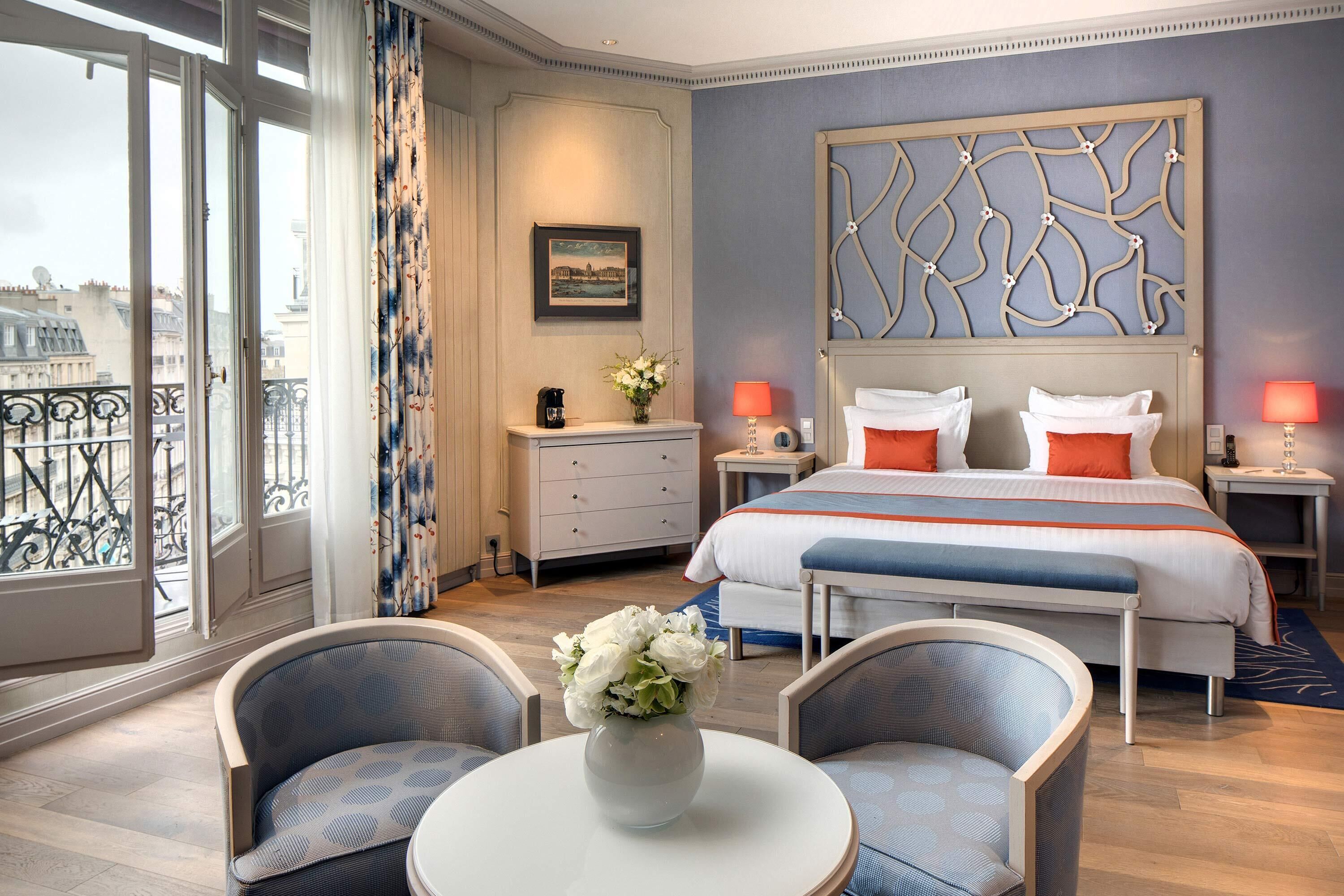 Habitación de hotel con una cama grande, dos mesitas de noche, una pequeña mesa con un jarrón de flores, dos sillas, un ventanal con vistas a edificios y un panel decorativo en la pared.