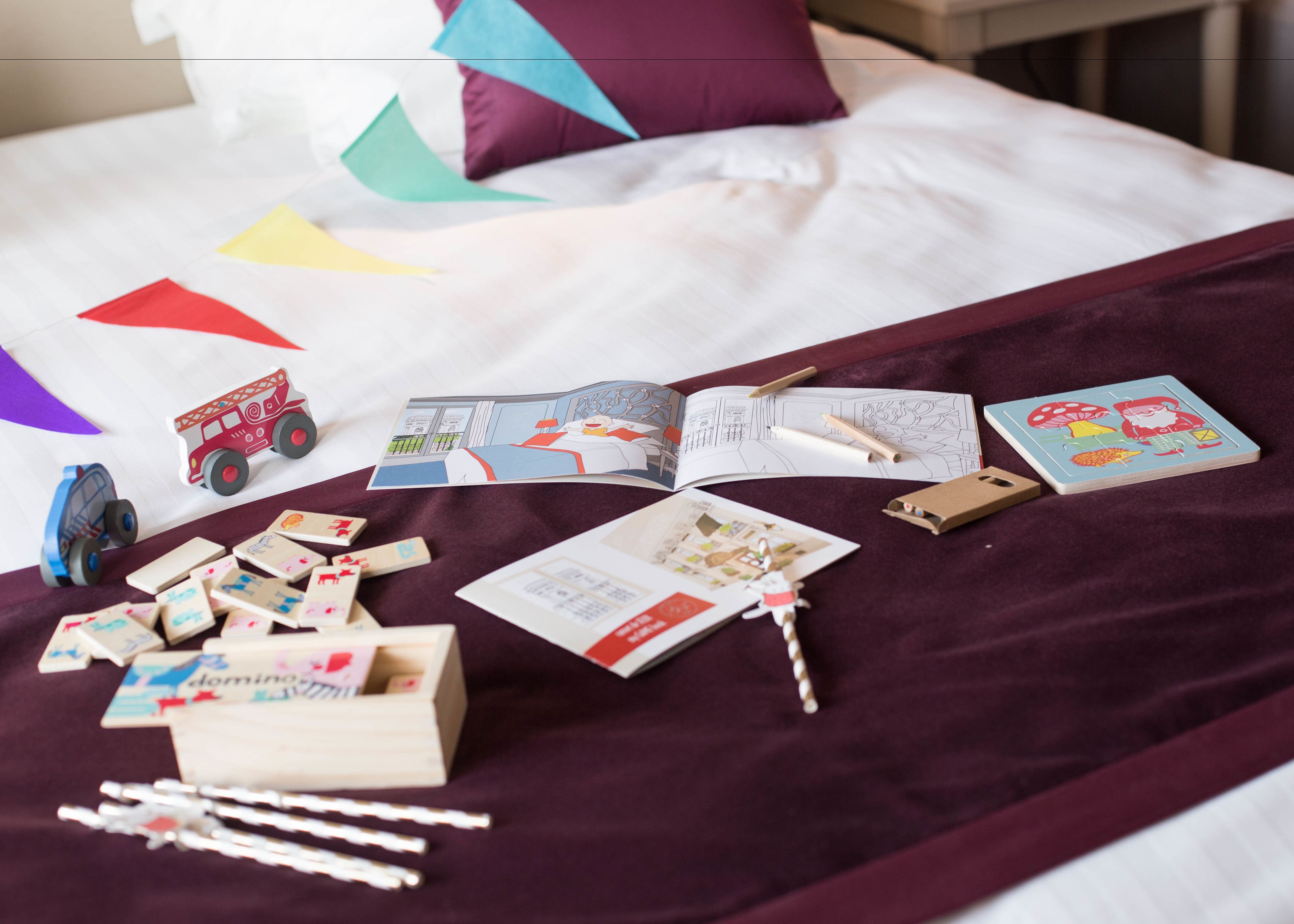 Ein Bett mit einer violetten Decke, einem Buch und verschiedenen Spielzeugen, darunter ein Feuerwehrwagen und Bauklötze.