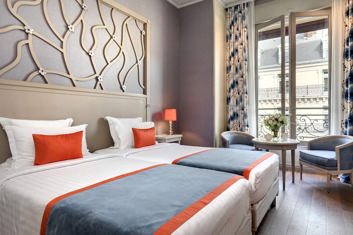 A hotel room with two beds, a nightstand with a lamp, a small table with a vase, and a chair.