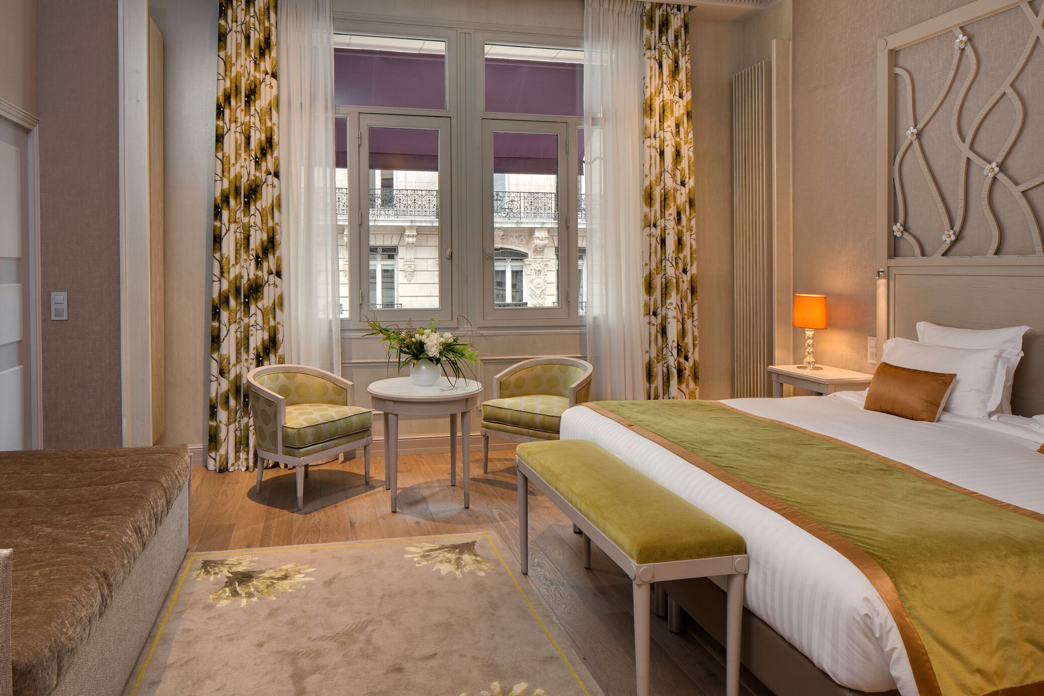 Habitación de hotel con una cama grande, una pequeña mesa redonda con sillas, una ventana con cortinas moradas y un cabecero decorativo.