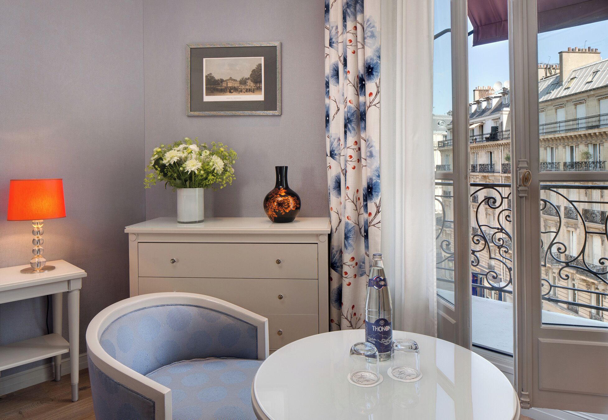 A room with a round table, a chair, a vase with flowers, a framed picture on the wall, and a view of buildings through a window.