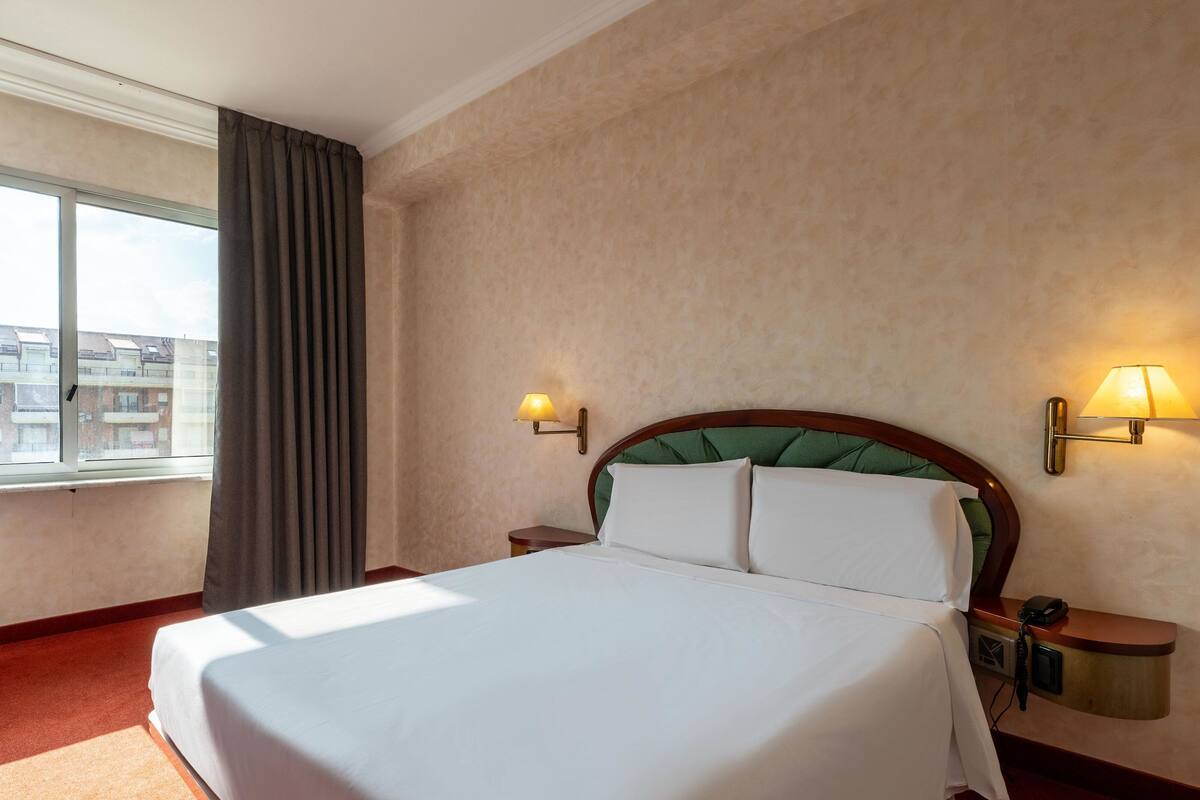 A hotel room with a large bed, two bedside tables, a nightstand, a window with curtains, and a view of buildings outside.