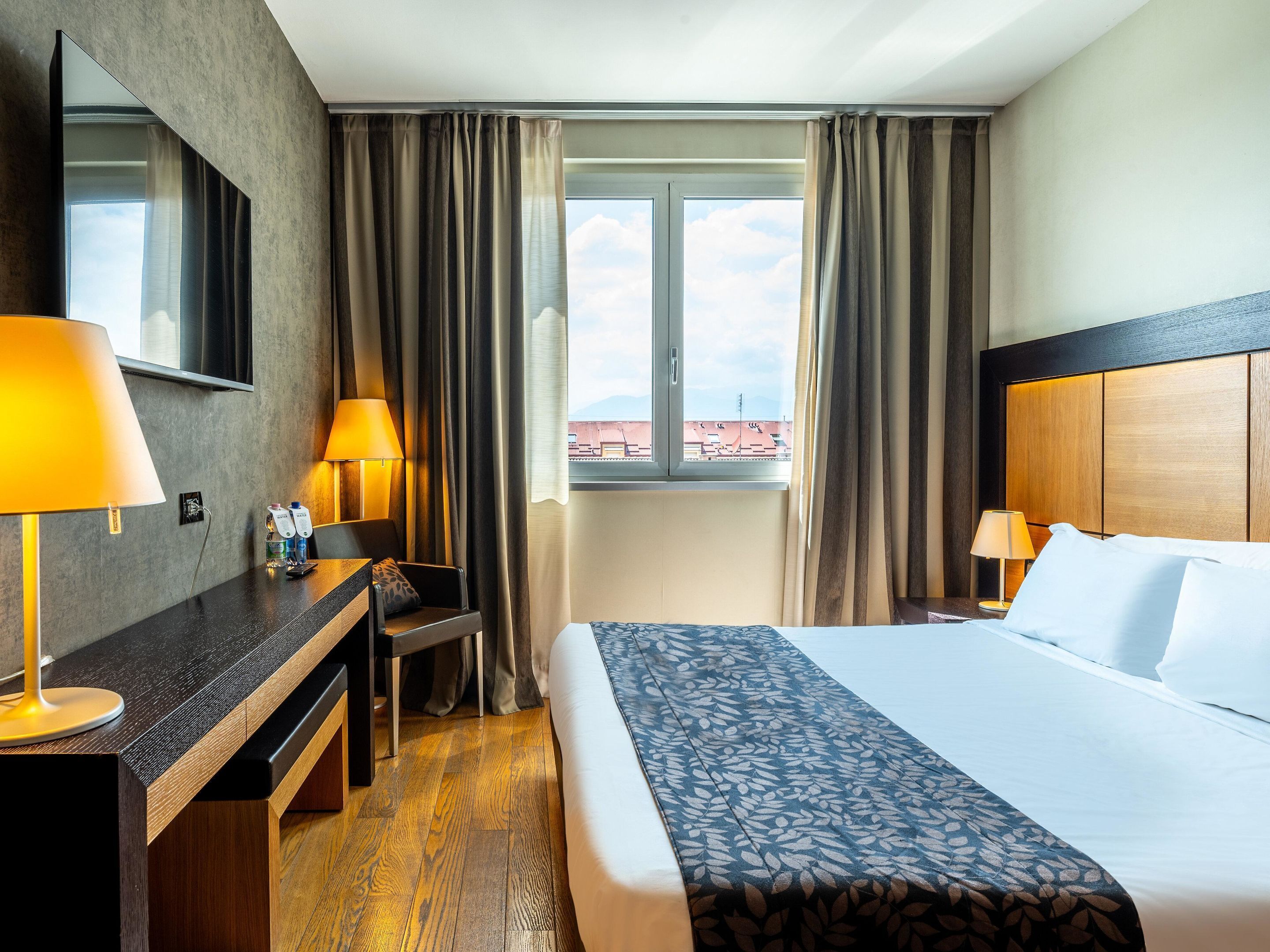 Une chambre d’hôtel comprenant un lit, deux lampes de chevet, un bureau avec une chaise, un grand miroir et une fenêtre avec des rideaux.
