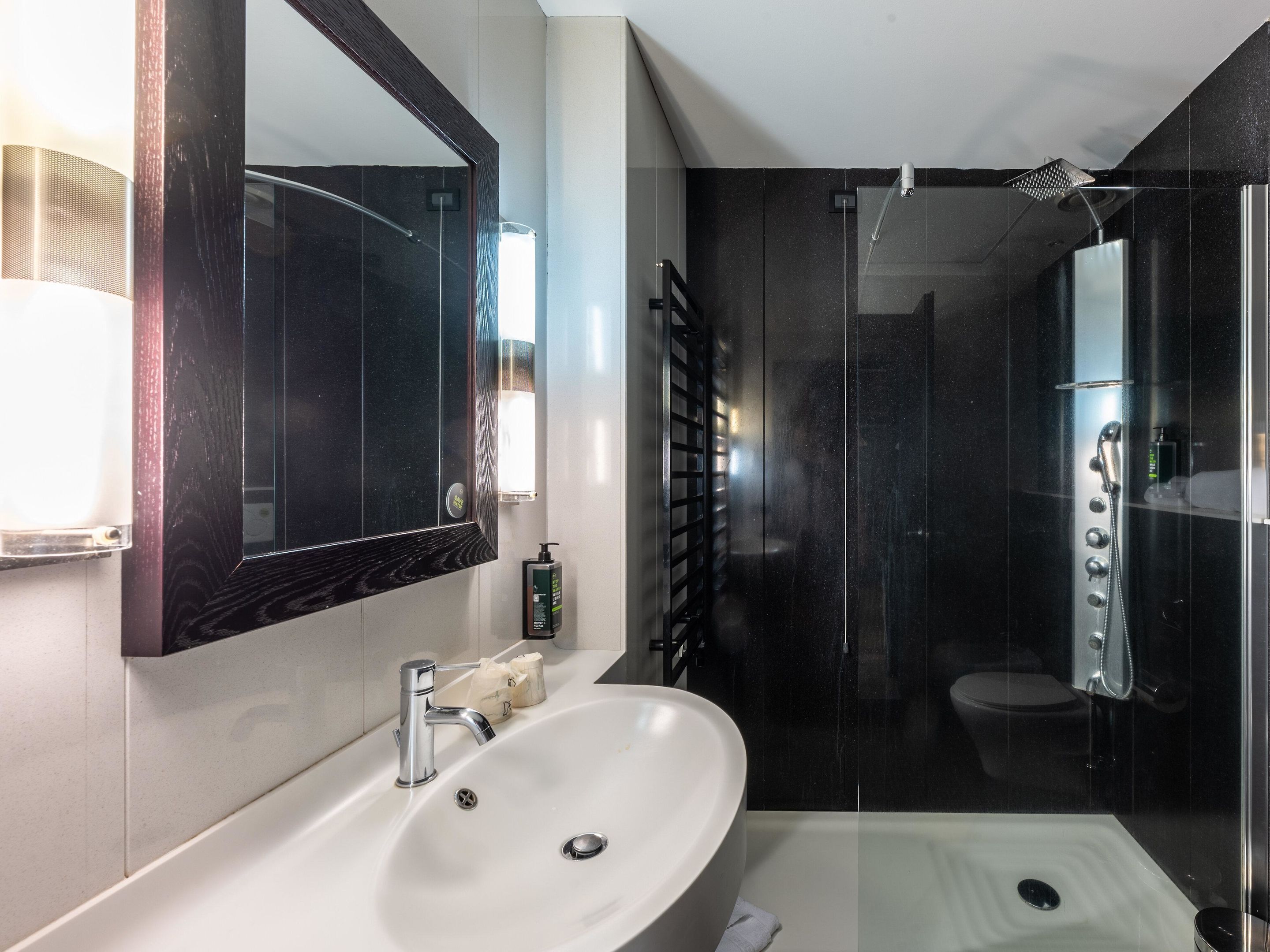 Un baño moderno con un espejo grande, un lavamanos blanco, una ducha con varios controles y un inodoro.