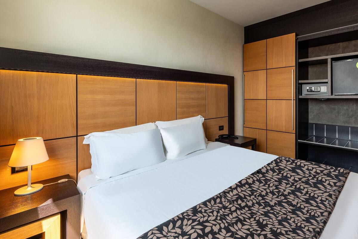 A hotel room with a large bed, a bedside table with a lamp, a nightstand, a built-in TV, and a minibar.