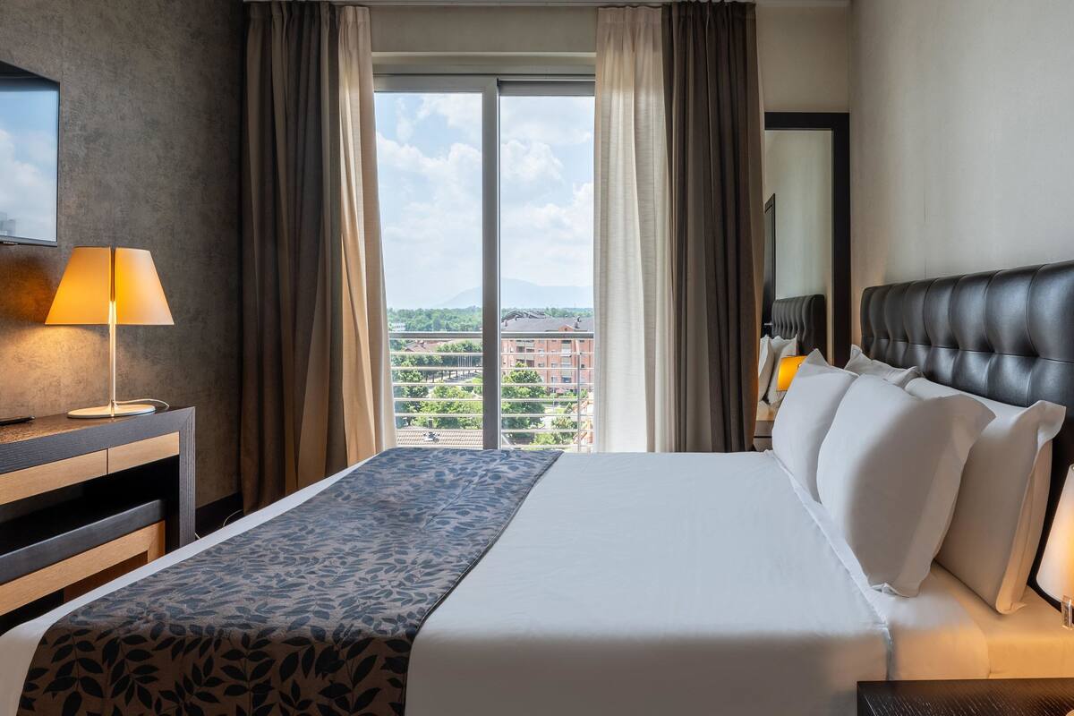 A hotel room with a large bed, a desk with a lamp, a view of the city through the window, and a cityscape visible outside.