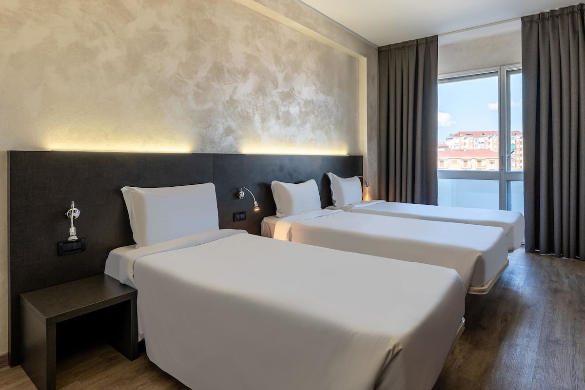 A hotel room with two beds, a nightstand, a wall-mounted lamp, and a large window with curtains.