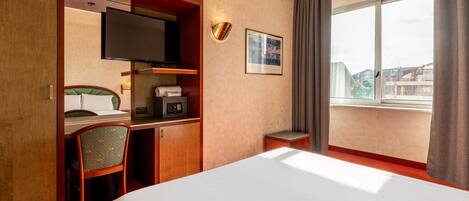 Camera d'albergo con un letto, una scrivania con un computer, una cassaforte, una TV e una finestra vista edifici.