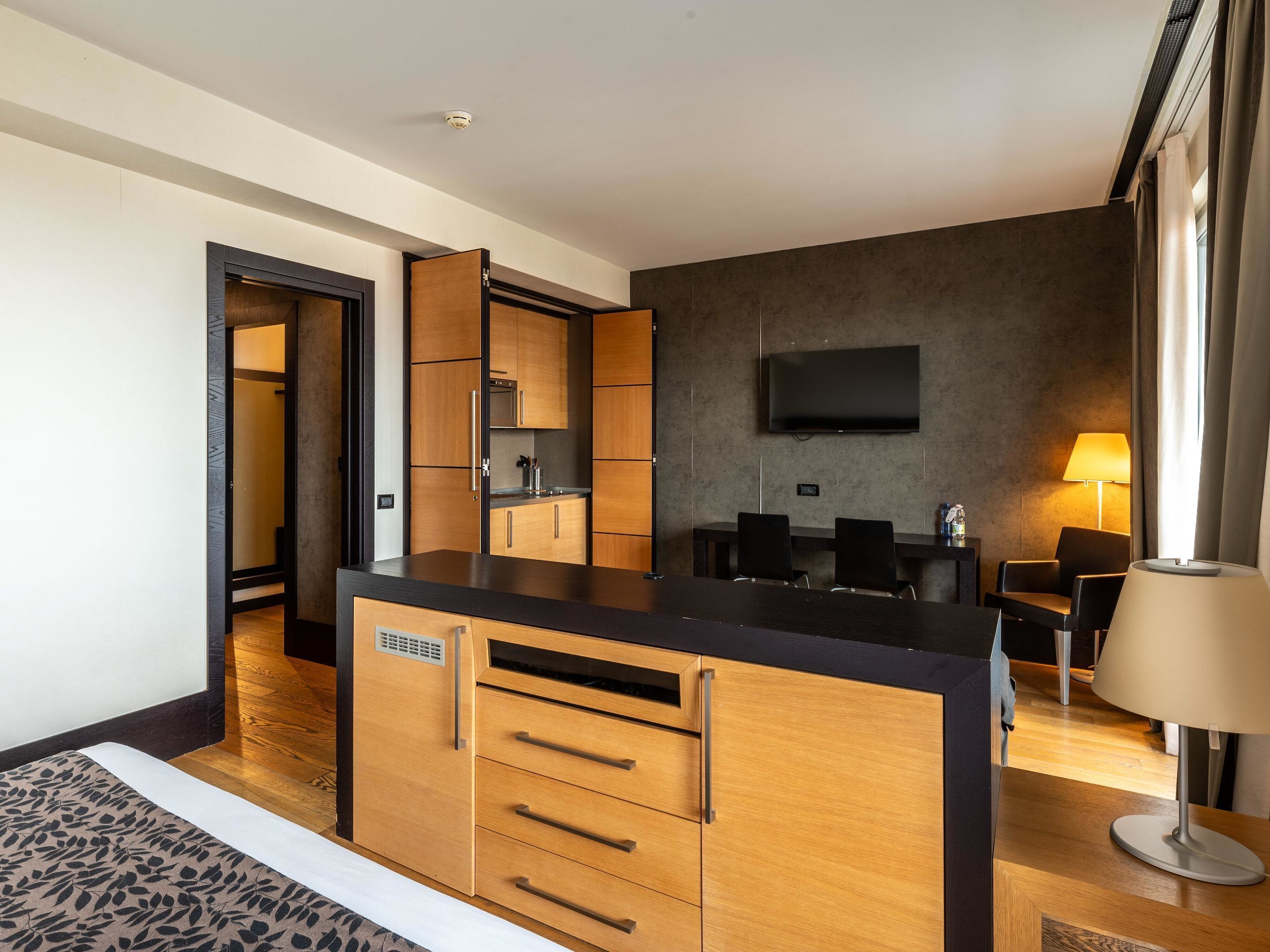 A modern hotel room with a wooden kitchenette, a flat-screen TV mounted on the wall, and a seating area with a table and chairs.